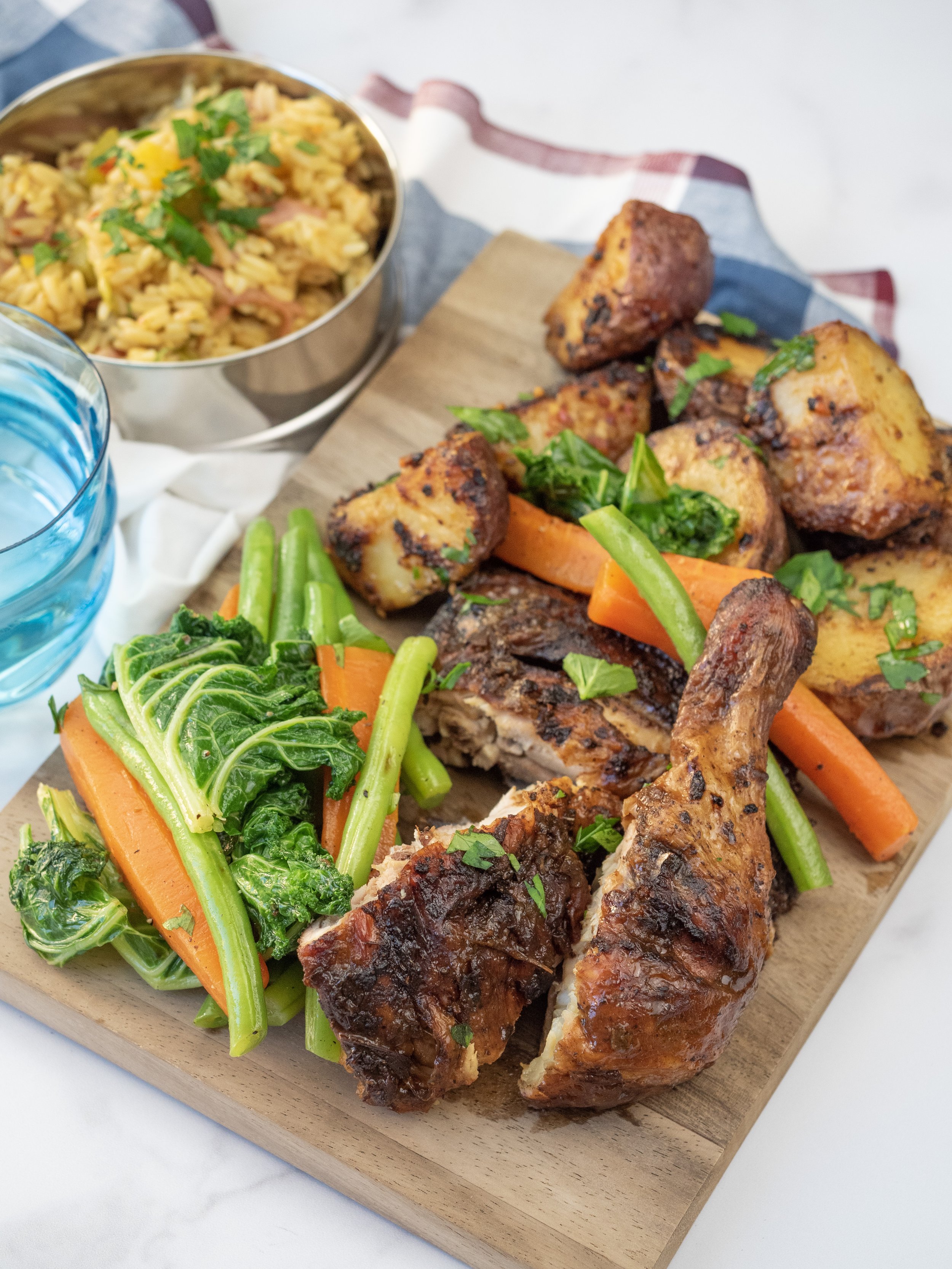 A wooden serving board with grilled chicken drumsticks, roasted potatoes, sautéed carrots, and greens, accompanied by bowls of rice and water.