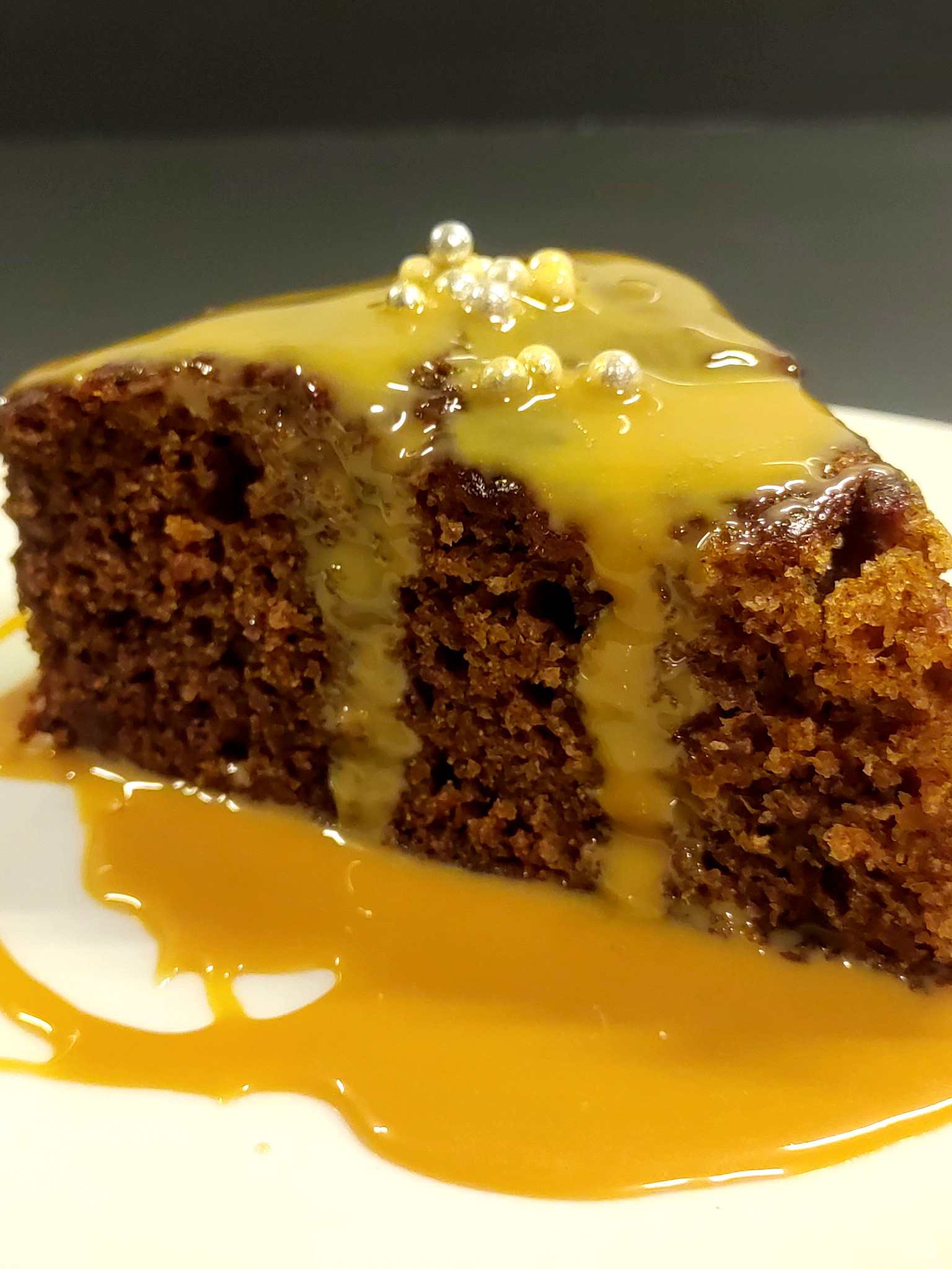 Close-up of a slice of chocolate cake with yellow caramel sauce and decorative sugar pearls on top, on a white plate.