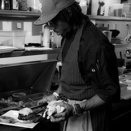 A chef wearing a striped apron, a hat, and glasses preparing food in a kitchen.
