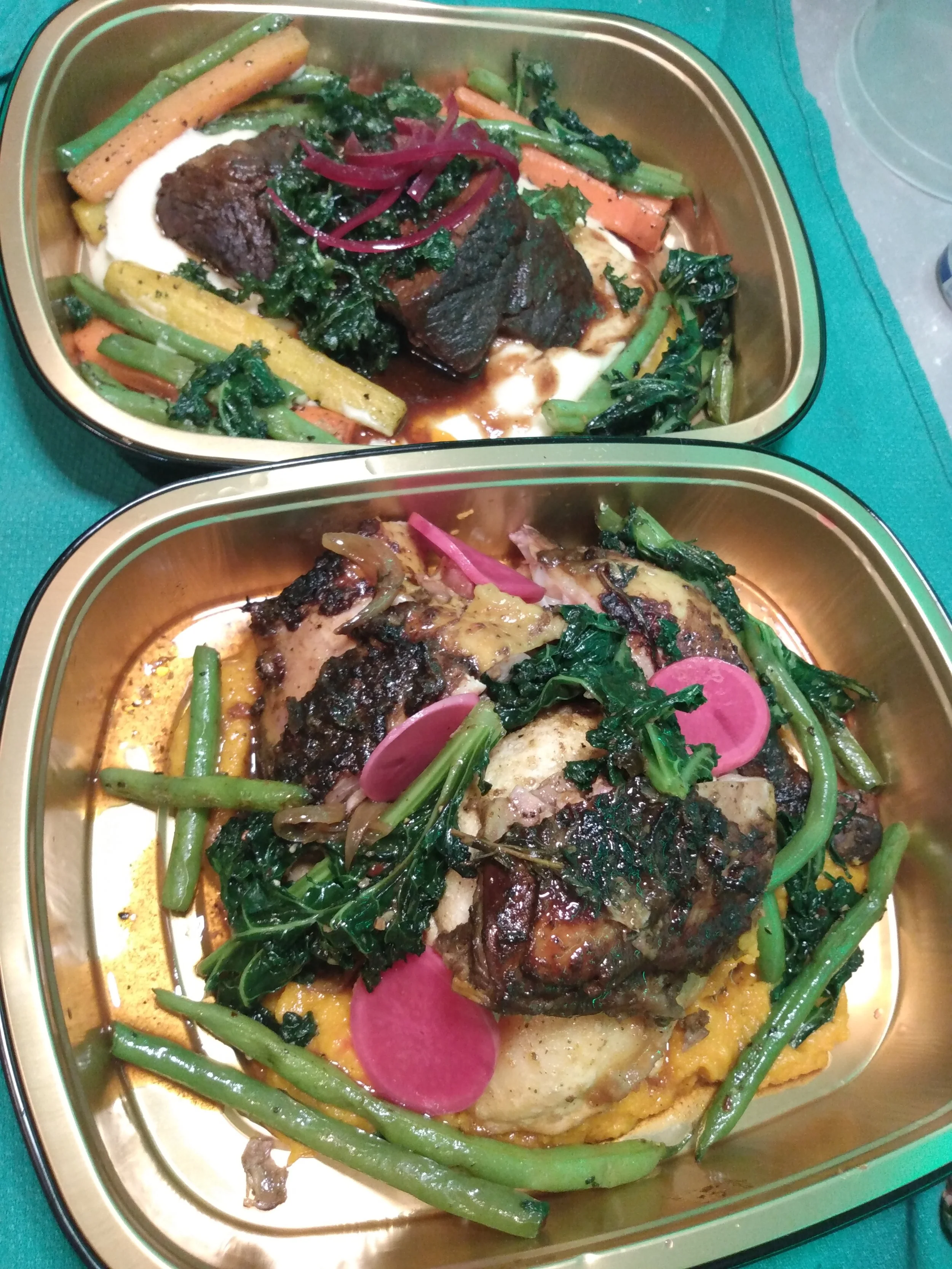 Two metal containers with cooked meat, green beans, carrots, kale, and pink radish slices on a teal table.