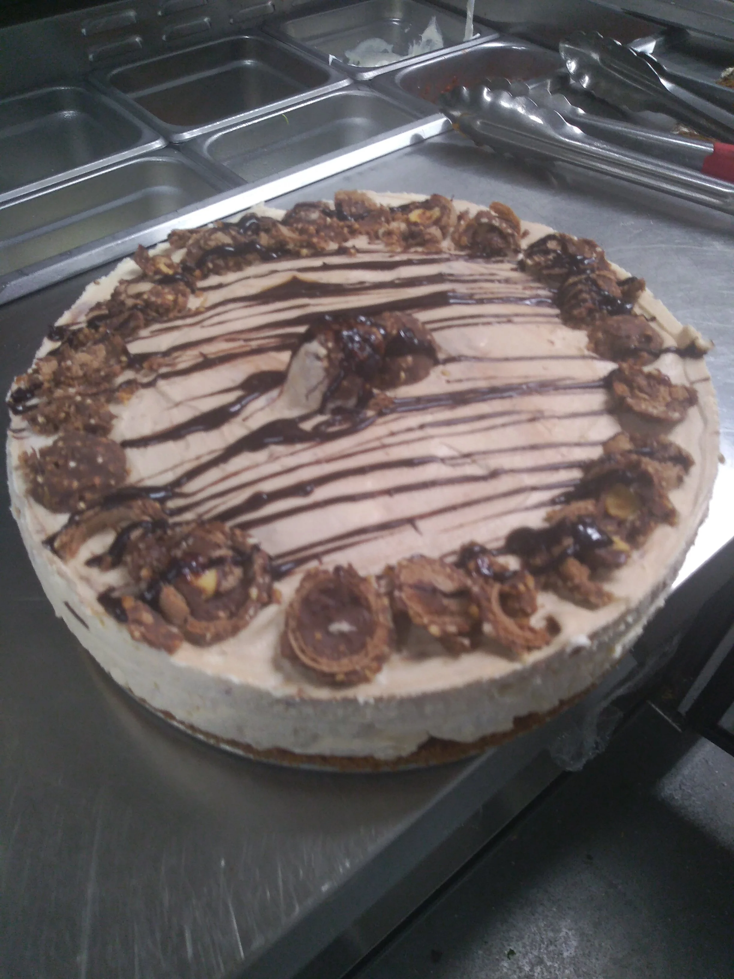 A large chocolate ice cream cake topped with chocolate drizzle and chocolate chip cookies around the edge, placed on a stainless steel counter.