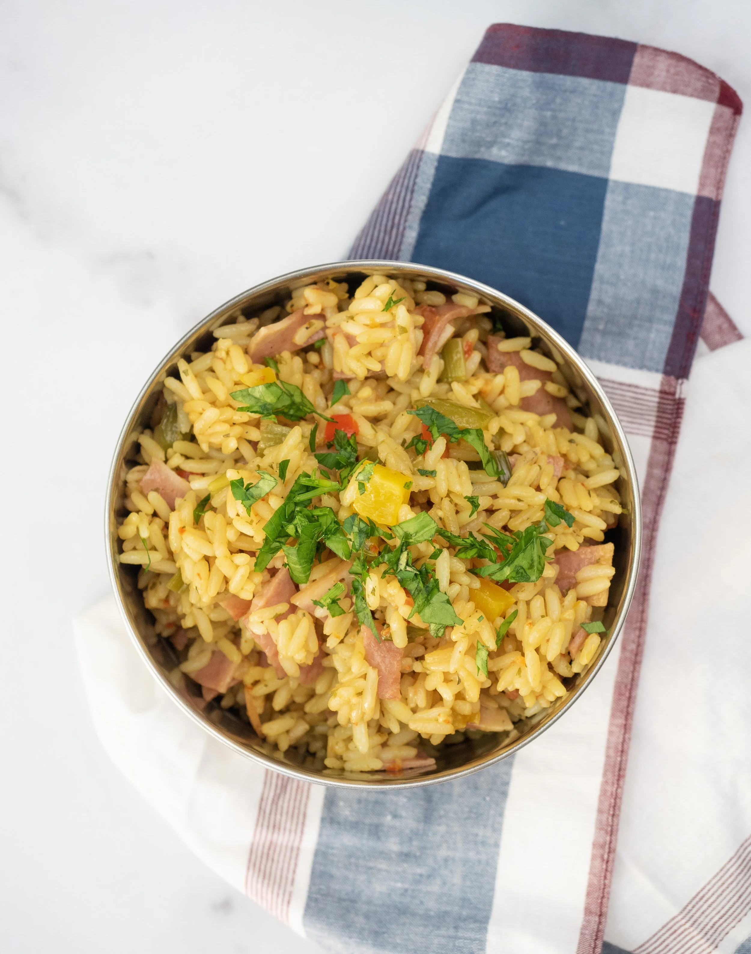A bowl of cooked rice mixed with vegetables and chopped herbs on a white surface with a folded checkered cloth napkin nearby.