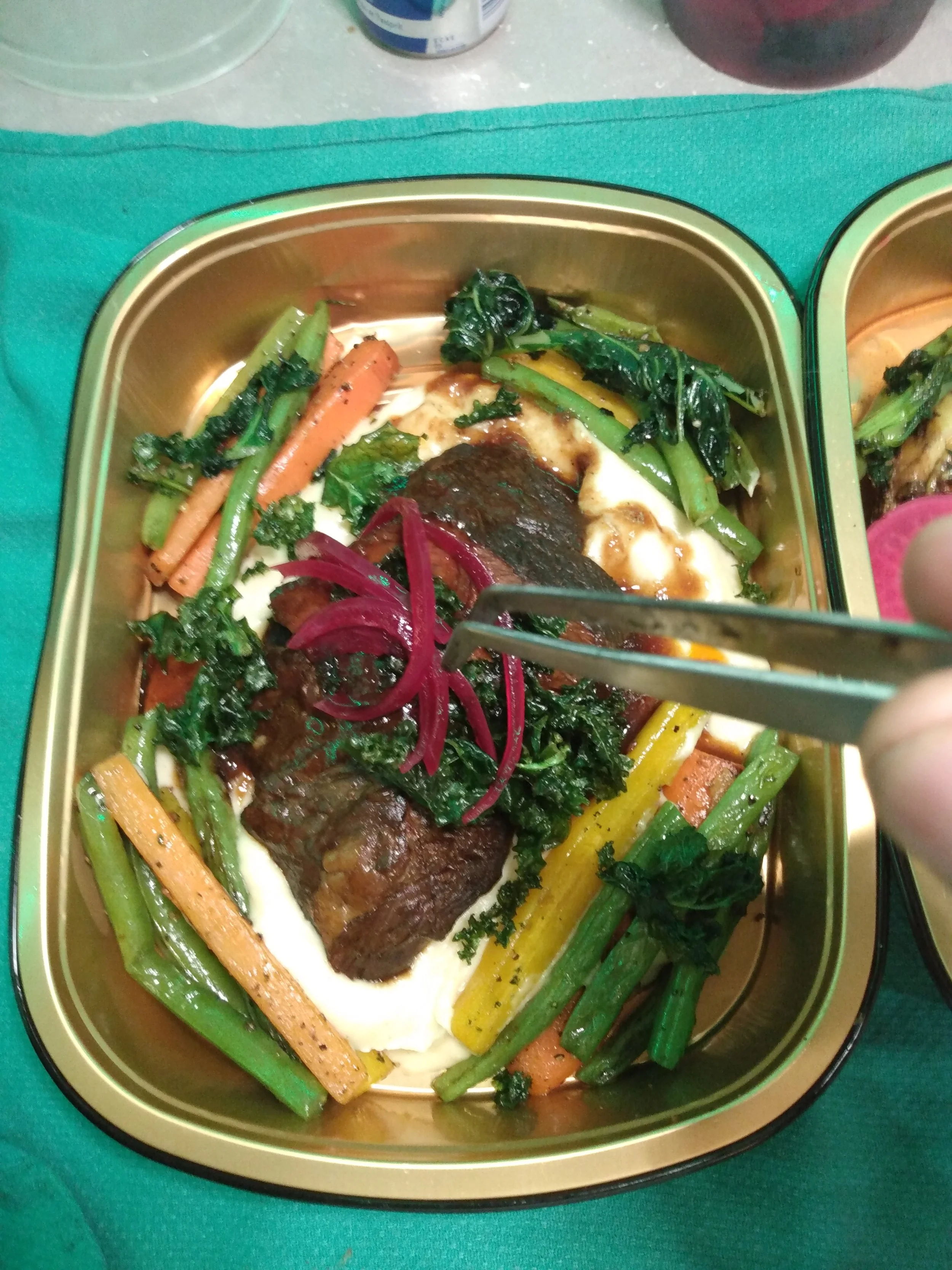 A tray of cooked vegetables, beef, and mashed potatoes, with a person using tongs to pick up some red onions.