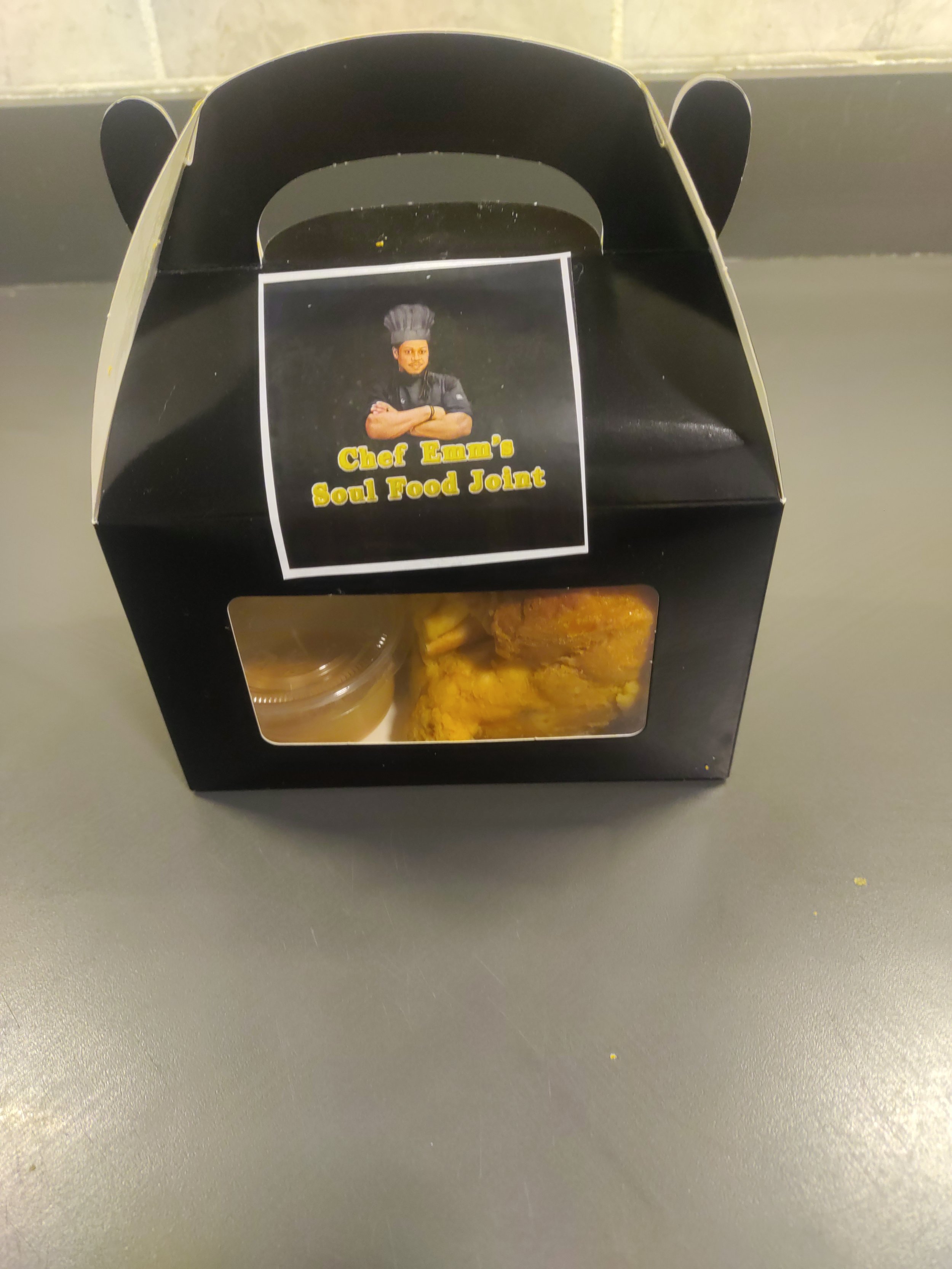 Black takeout box with a label that features a picture of a chef in a black uniform and hat, with arms crossed, and the text 'Chef Emi's Soul Food Joint'. The box contains food, with a small container of gravy and a piece of fried chicken visible thr