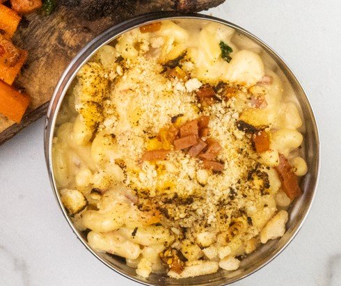 A bowl of baked macaroni and cheese topped with breadcrumbs, ham, and herbs