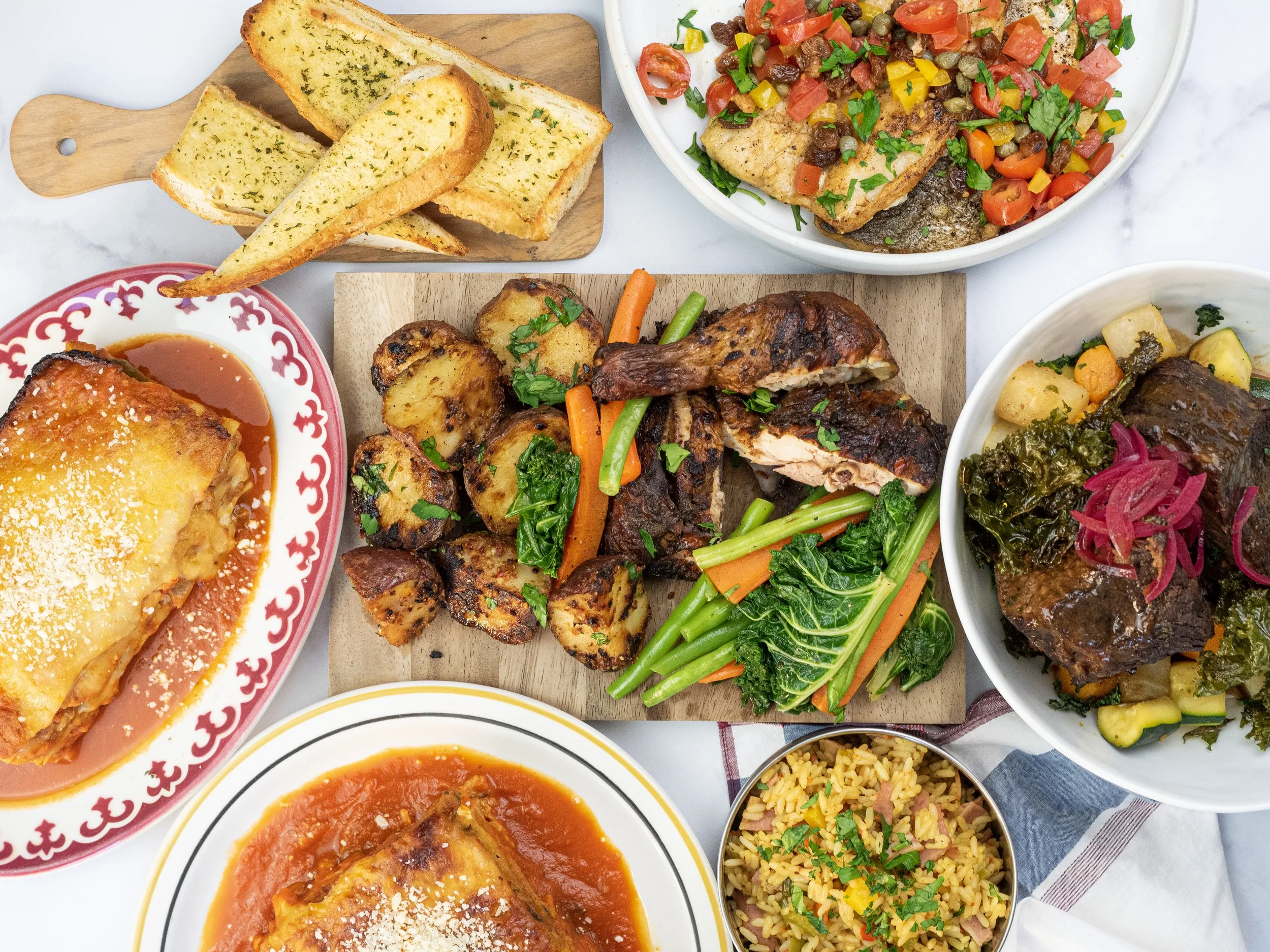 A variety of foods on a table including garlic bread, baklava, roasted vegetables with chicken, a meat and potato casserole, rice, and a vegetable stew with beef.
