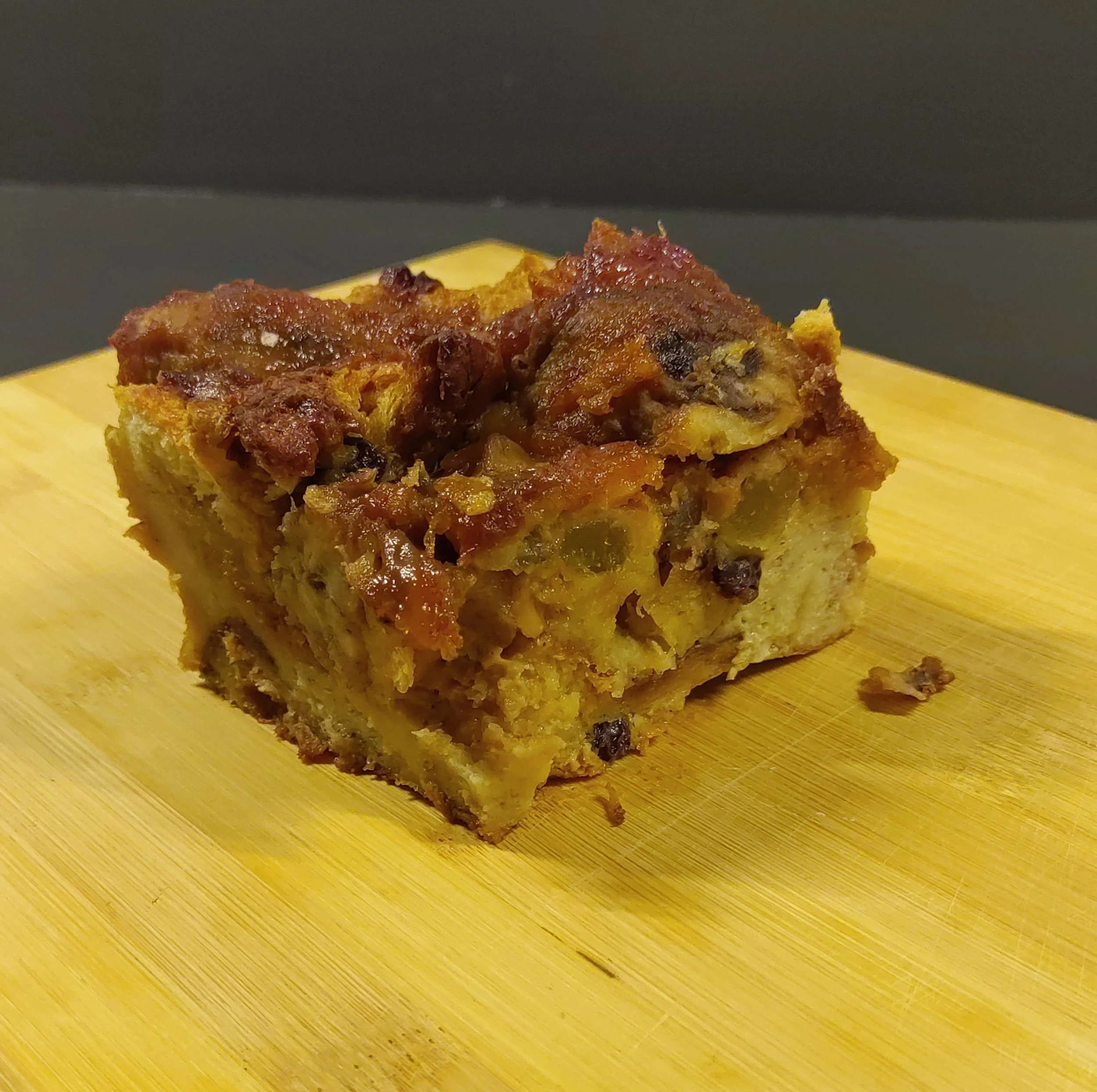 A close-up of a slice of homemade baked bread pudding with caramelized bananas, and raisins, on a wooden cutting board.