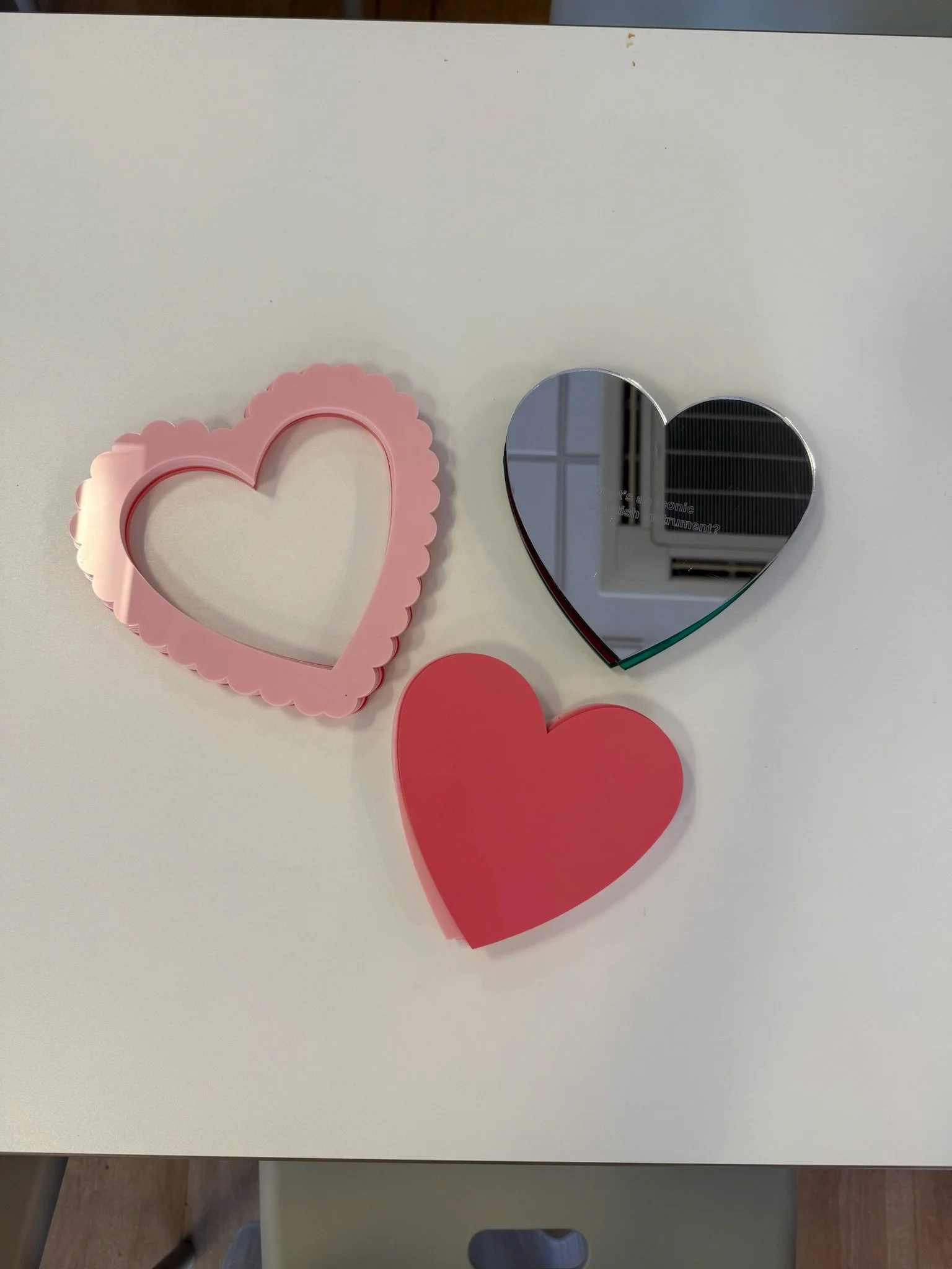 A pink heart-shaped photo frame, a silver heart-shaped mirror, and a red paper heart on a white table.