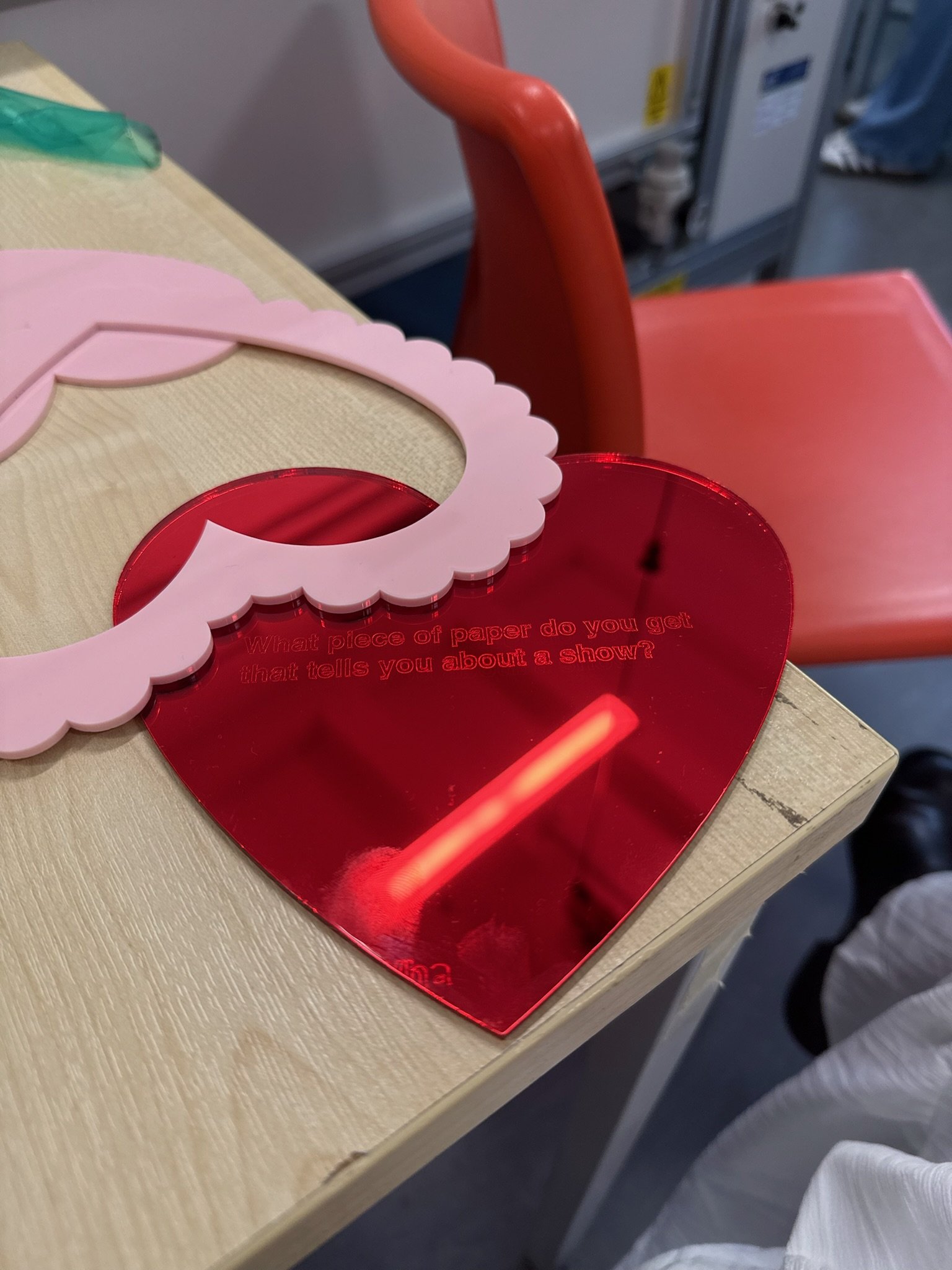 Close-up image of a pink valentine's day card with a scalloped edge, a red heart-shaped acrylic piece with question text, and a wooden surface. The red heart-shaped piece has the text 'What piece of paper do you get that tells you about a show?' engraved on it.