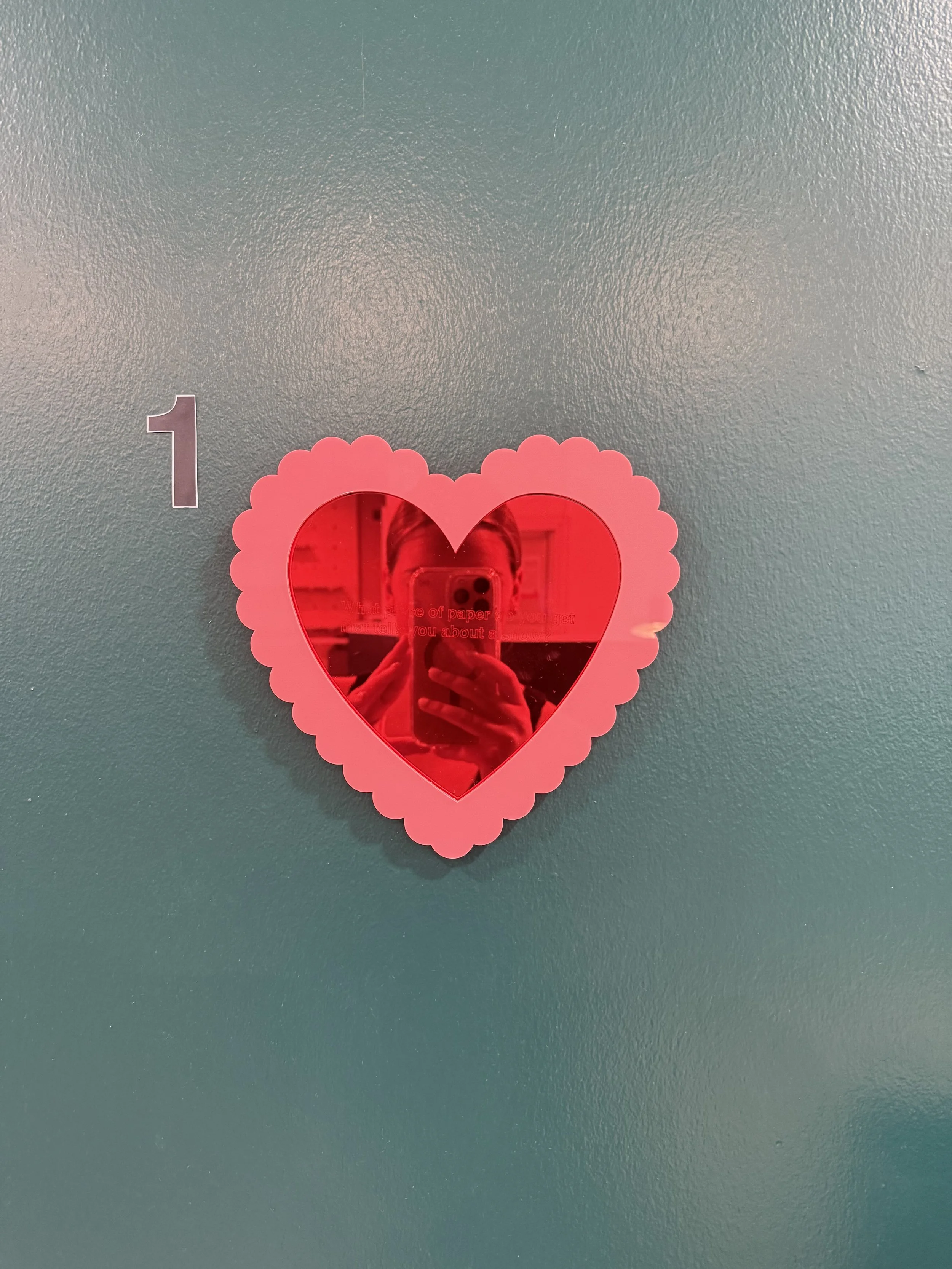 A pink heart-shaped mirror with a scalloped border hanging on a teal wall, reflecting a person taking a photo with a smartphone.
