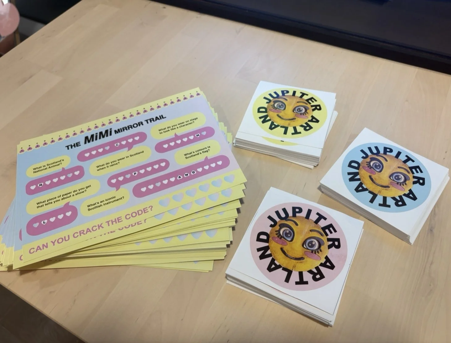Colorful quiz sheets titled 'The MiMi Mirror Trail' with questions about Scotland, and emoji emoticons, alongside stacks of cards featuring a smiling, winking cartoon face with the words 'Land JuPITER AR' in a circle.