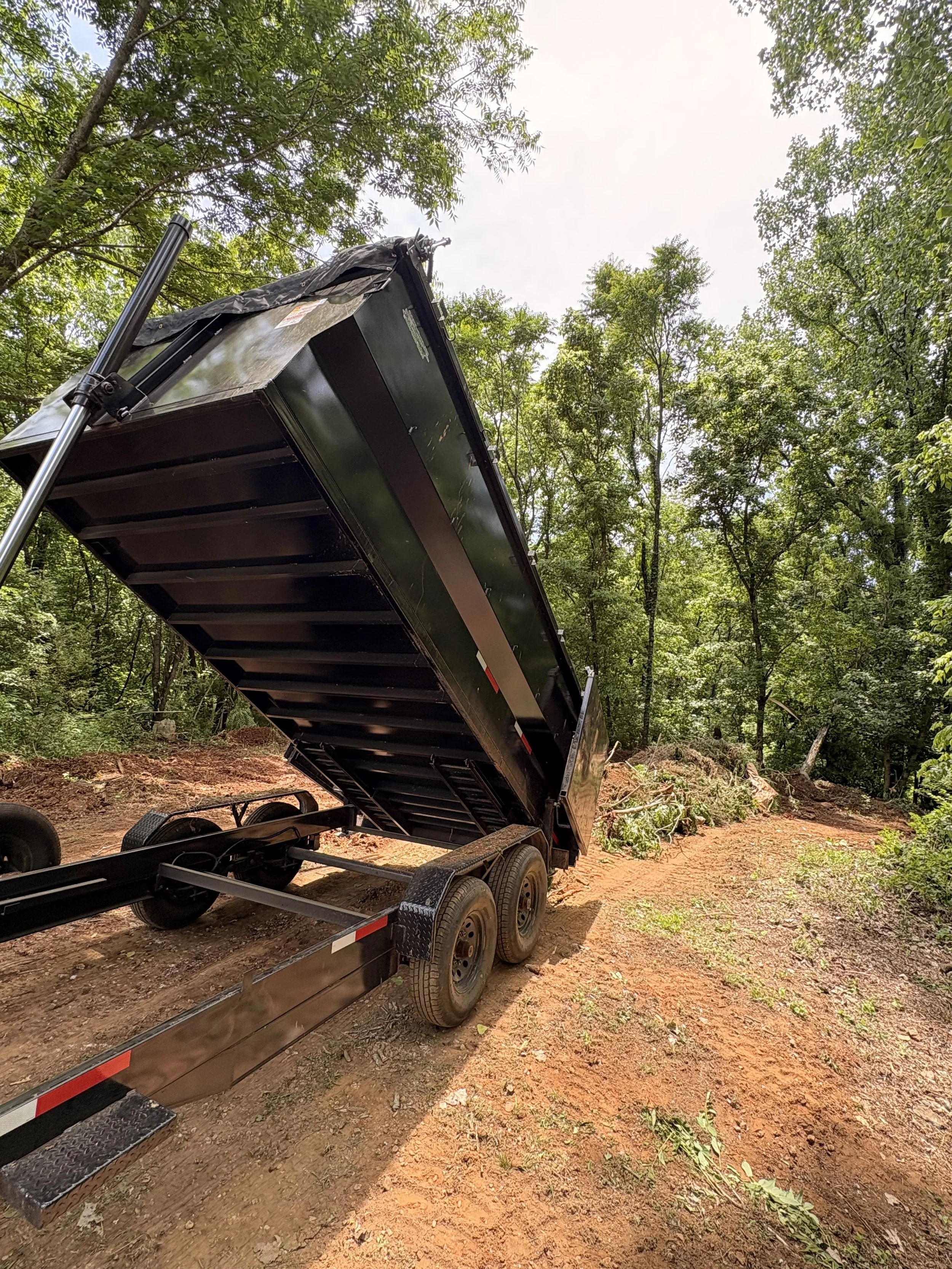 A black dump trailer with its bed raised, parked on a dirt path surrounded by trees in a forested area. Storm Cleanup, Debris Removal