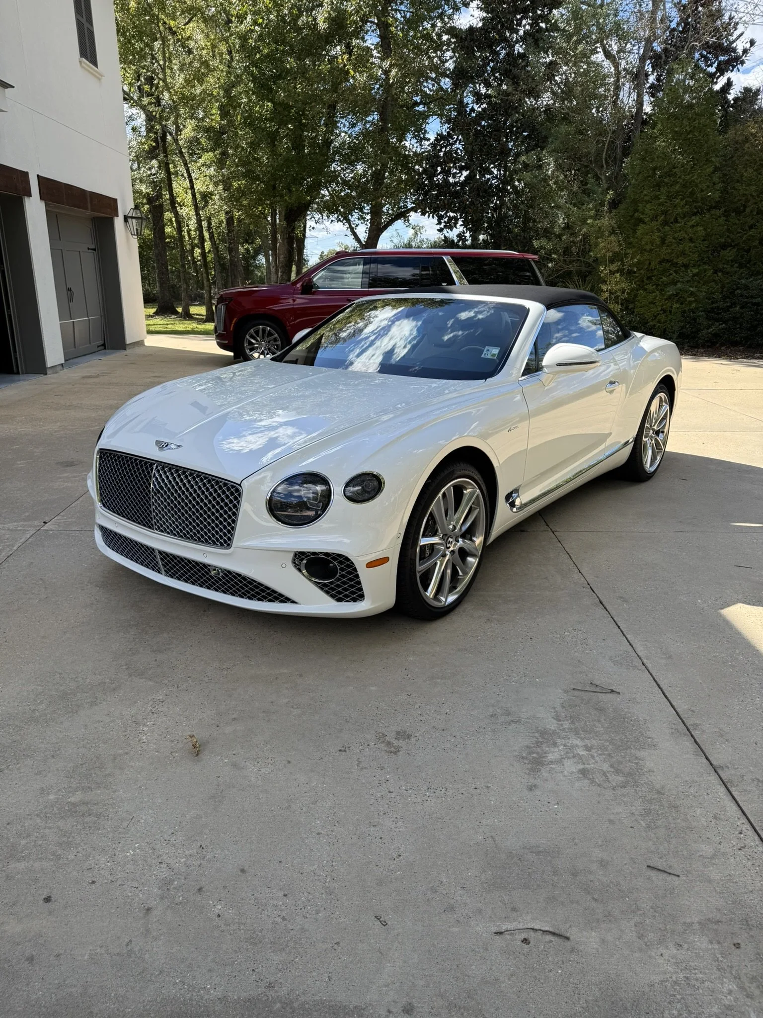 A white luxury convertible car parked on a driveway with a red SUV in the background and trees surrounding the area.
