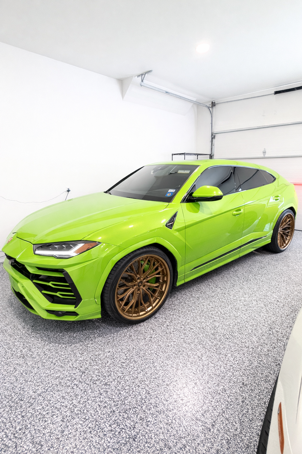 A bright green modern car with bronze wheels parked inside a garage with white walls and gray speckled flooring.
