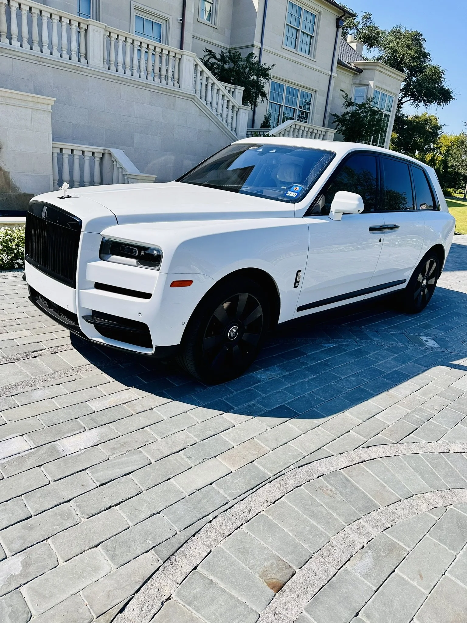 White luxury SUV, possibly a Rolls-Royce, parked on a paved driveway in front of a large, elegant house with stone walls and stairs, under a clear blue sky.