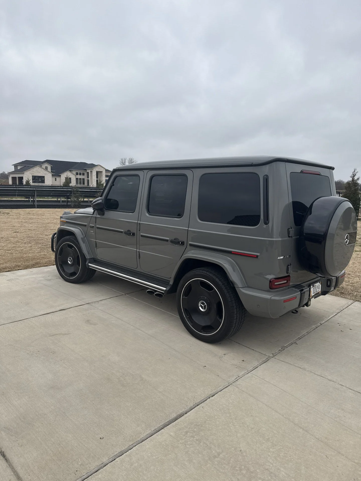 Gray Mercedes-Benz G-Class SUV parked on a concrete driveway in a suburban neighborhood, with nearby houses and overcast sky.