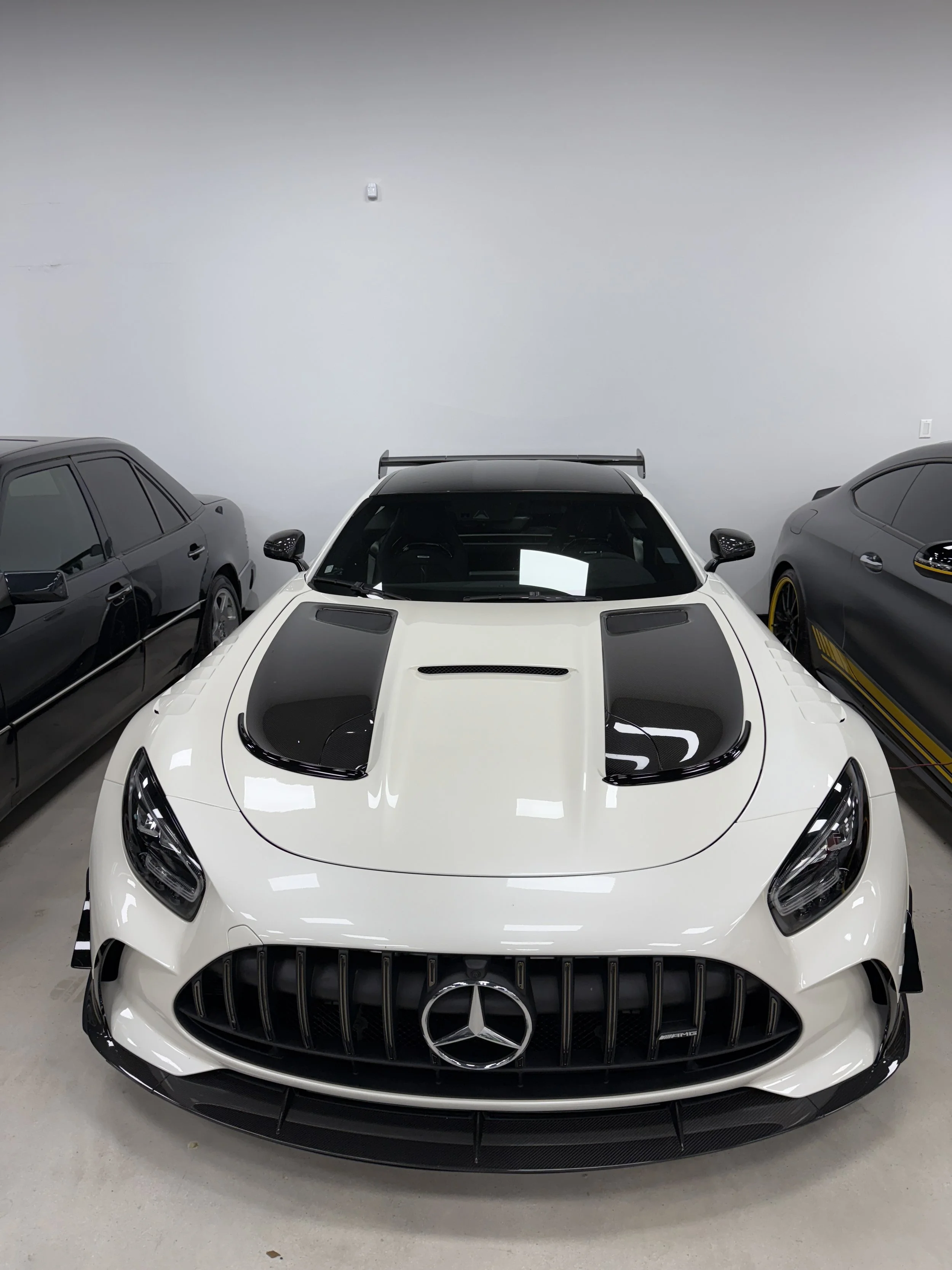 A white Mercedes-Benz sports car with black accents and a large Mercedes emblem on the front grille, parked indoors between two other high-end cars.