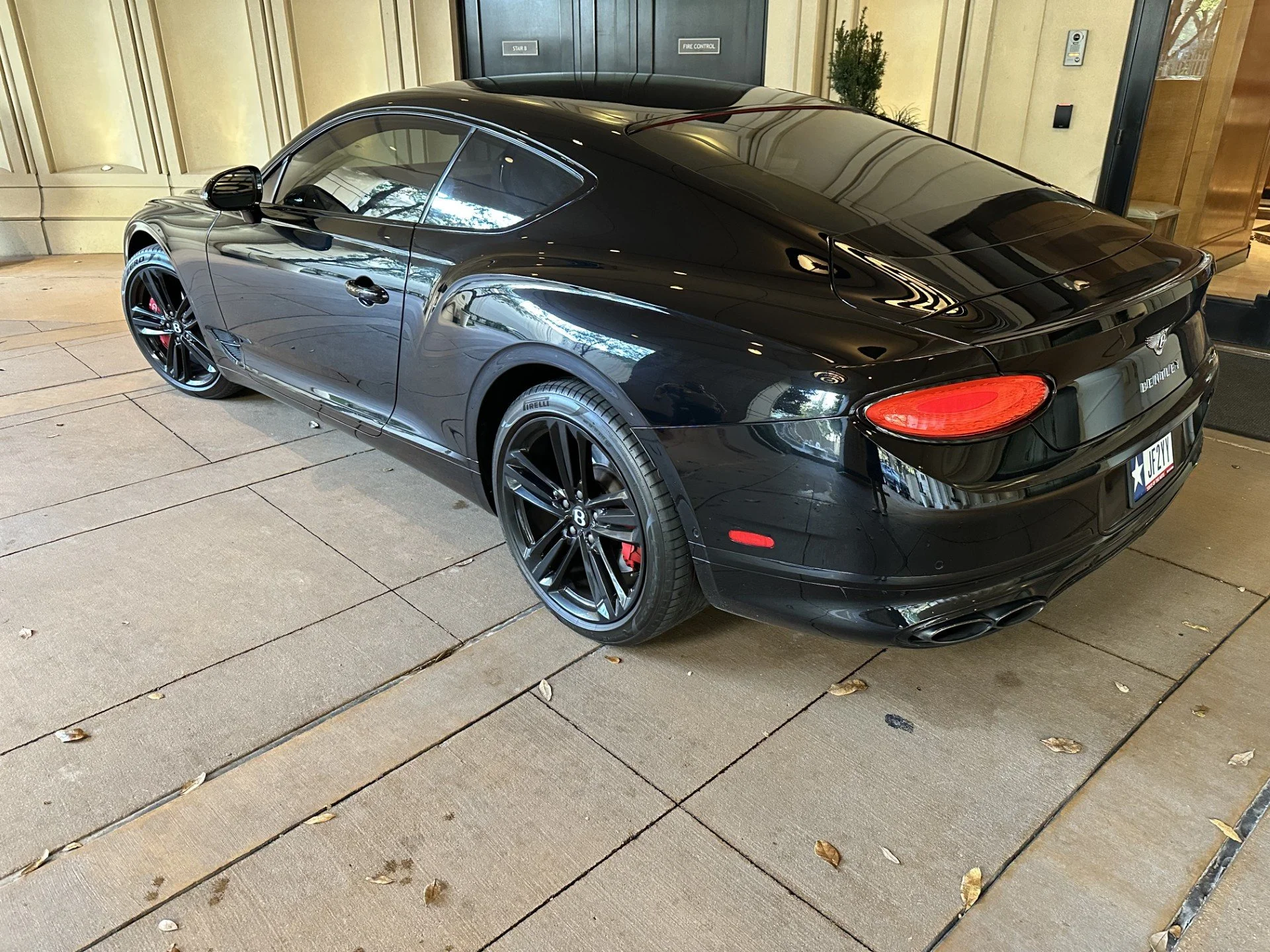 Black Bentley Continental GT parked on a paved sidewalk outside a building with beige stone walls and glass doors.