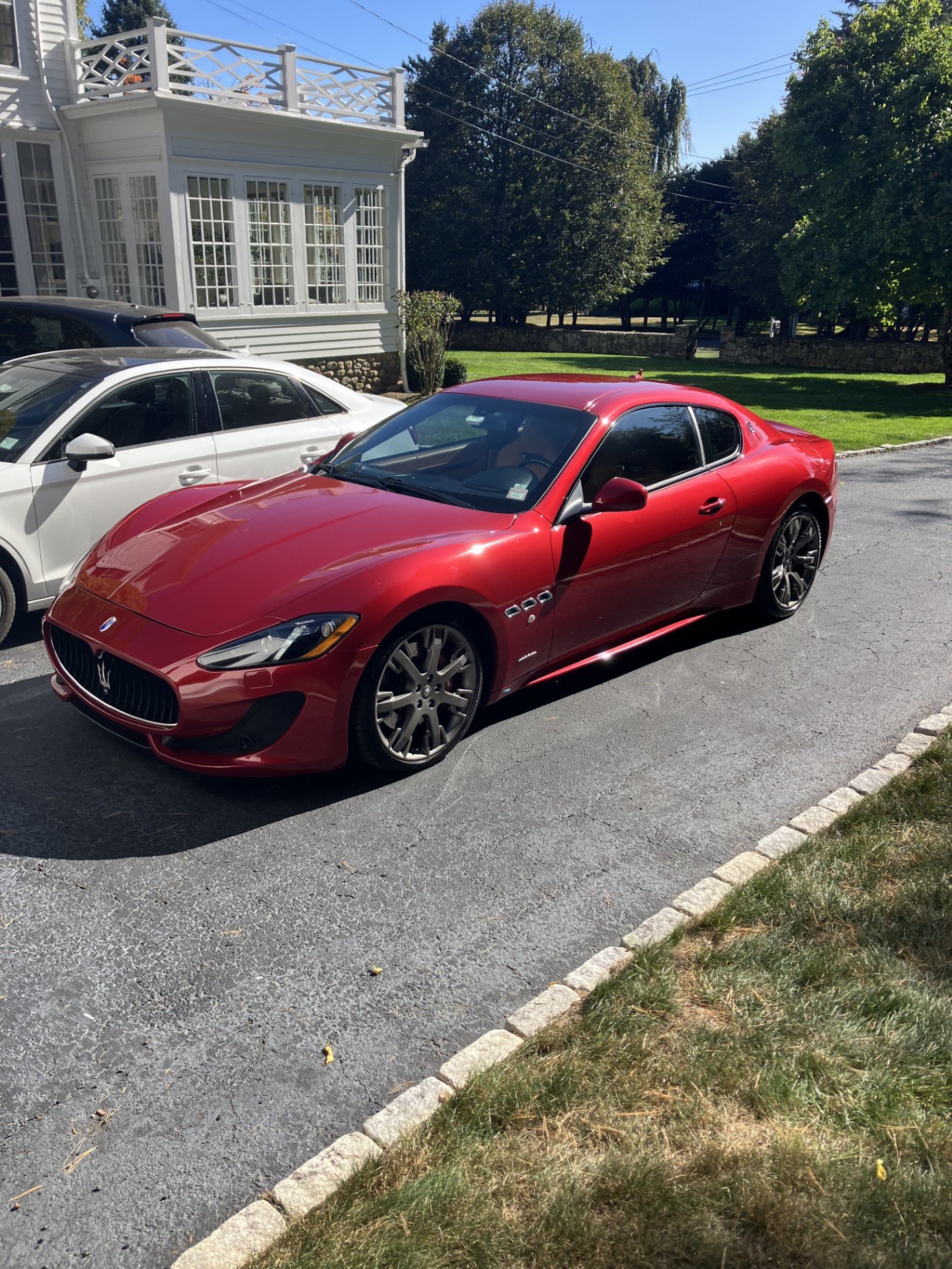 A red Maserati GranTurismo sports car parked on a driveway next to a white car, with a white house and trees in the background under a clear blue sky.