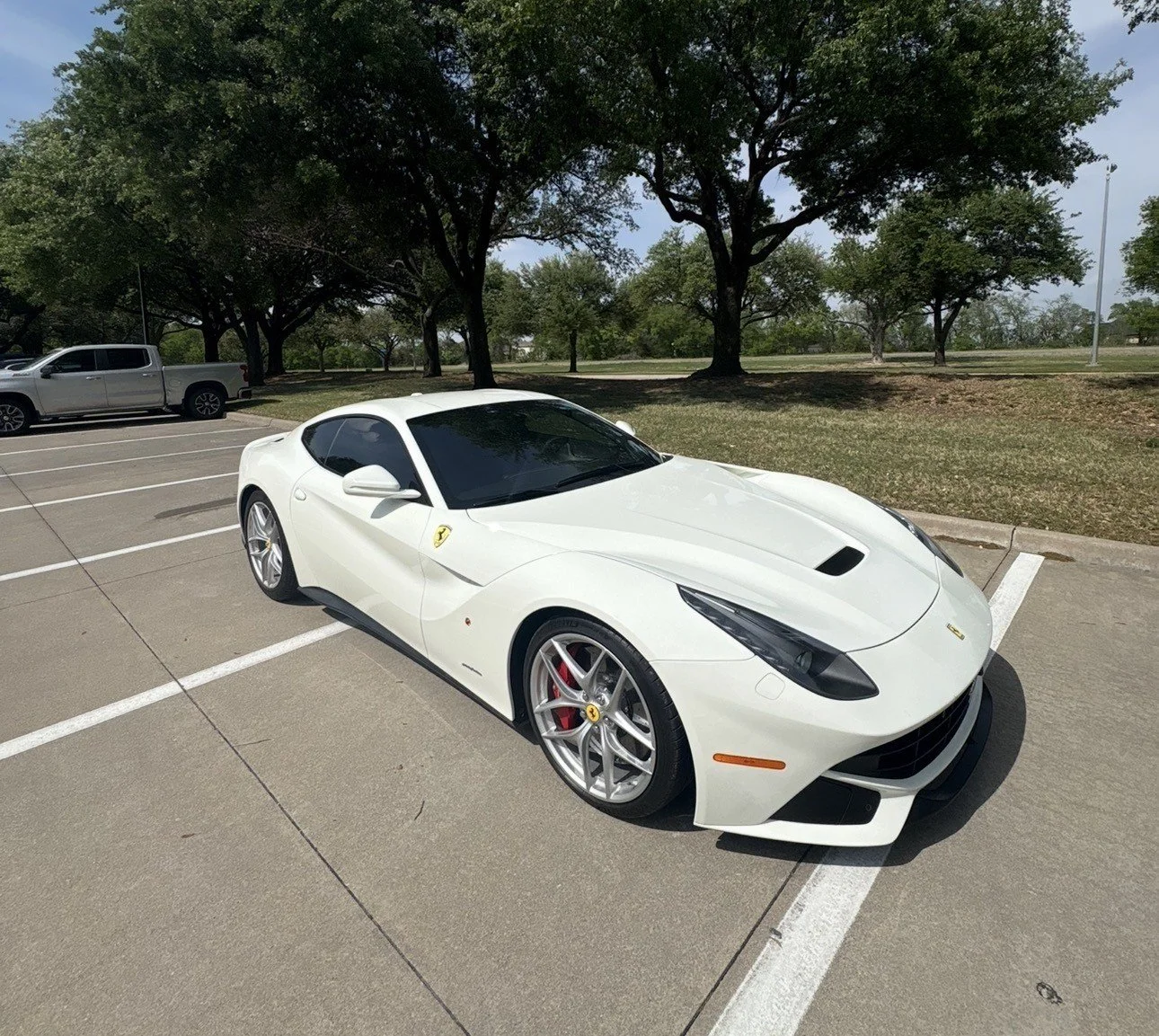 A white Ferrari sports car parked in a parking lot with trees and a grassy area in the background.