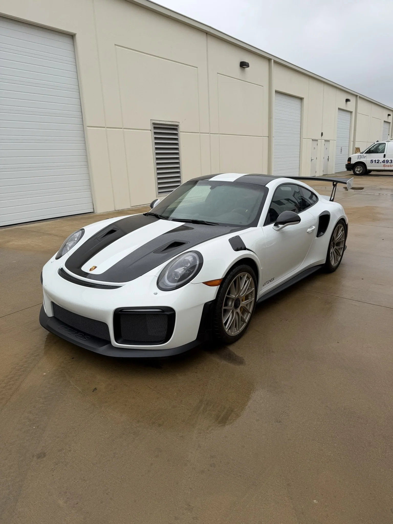 A white Porsche 718 Cayman GT4 sports car with black racing stripes parked outside a beige industrial building on a wet concrete surface.