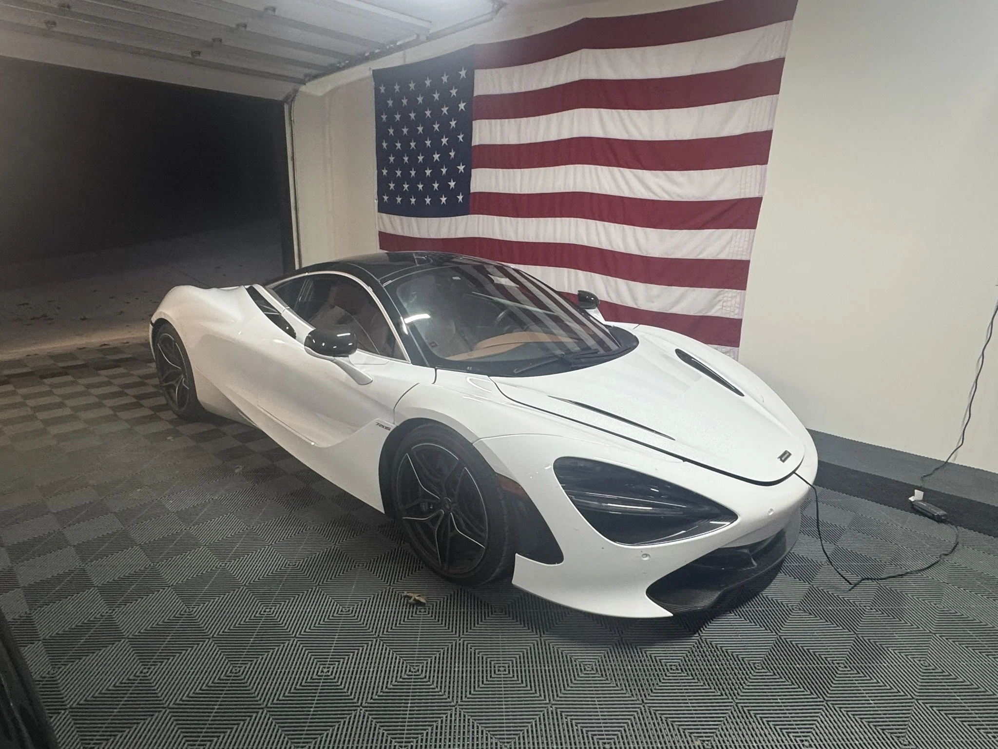 A white sports car with black accents parked in a garage, American flag hanging on the wall behind it.
