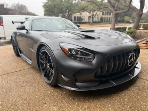 Black Mercedes-Benz sports car with a matte finish, large front grille, and sleek design parked on a driveway with trees and a house in the background.