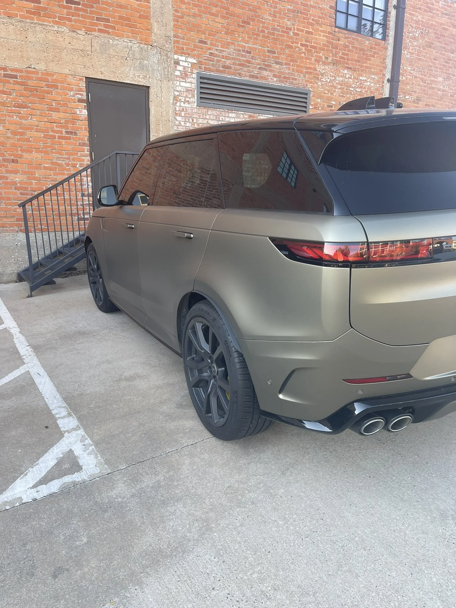 A beige luxury SUV with dark tinted windows parked in an outdoor lot near a brick building. The vehicle features black multi-spoke wheels, quad exhaust pipes, and an angular design.