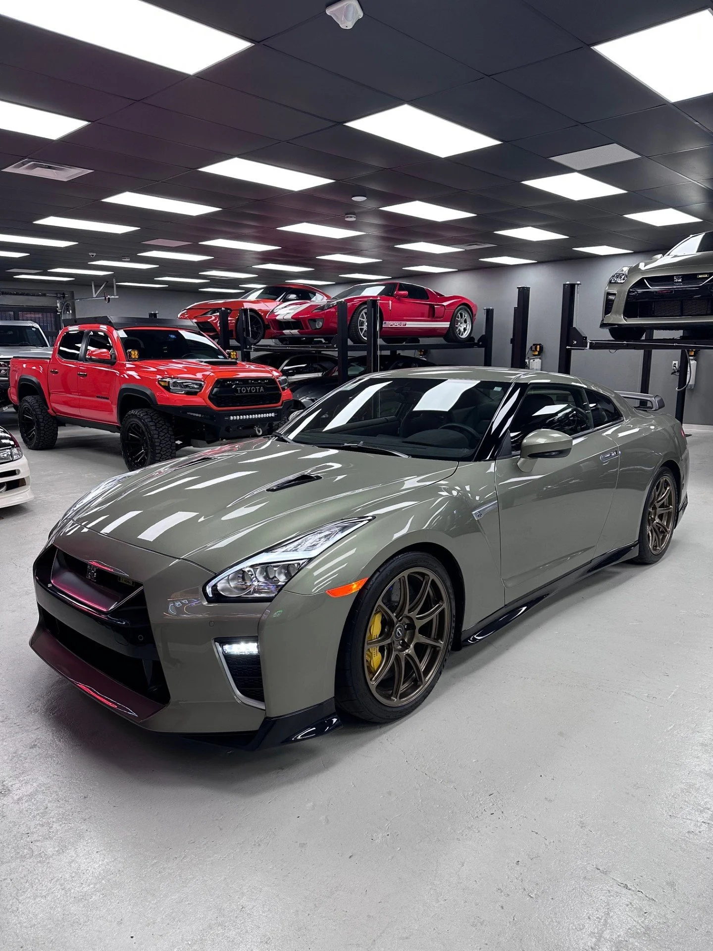 A gray Nissan GT-R sports car in a garage with other luxury and off-road vehicles, including a red Toyota truck, a red sports car on a lift, and a silver car on an upper lift.
