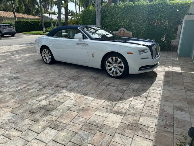 White luxury convertible car parked on a paved driveway with trees and bushes in the background.