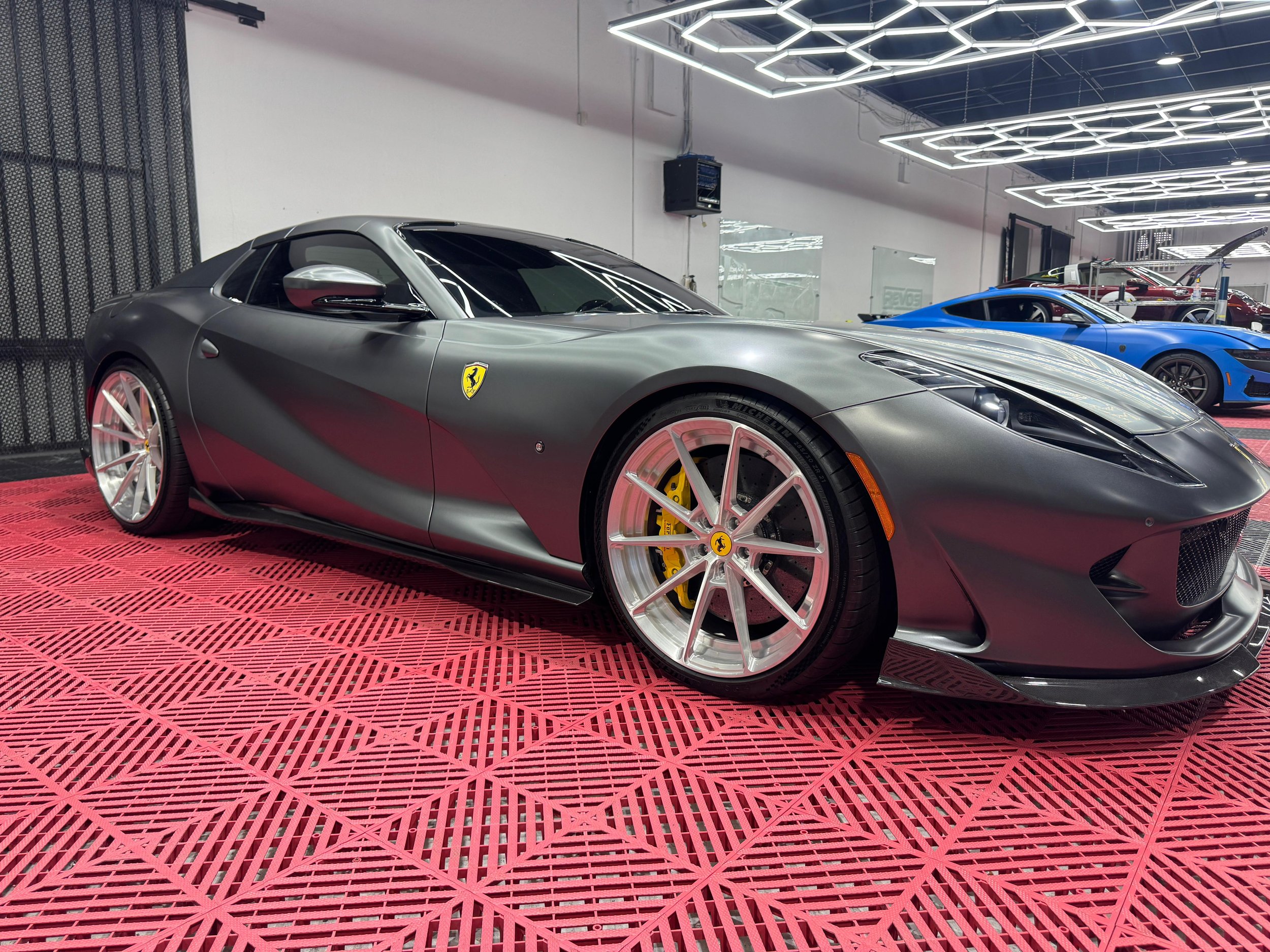 A metallic gray Ferrari sports car on a red platform inside a modern showroom with bright ceiling lights, parked next to a blue sports car and other vehicles.