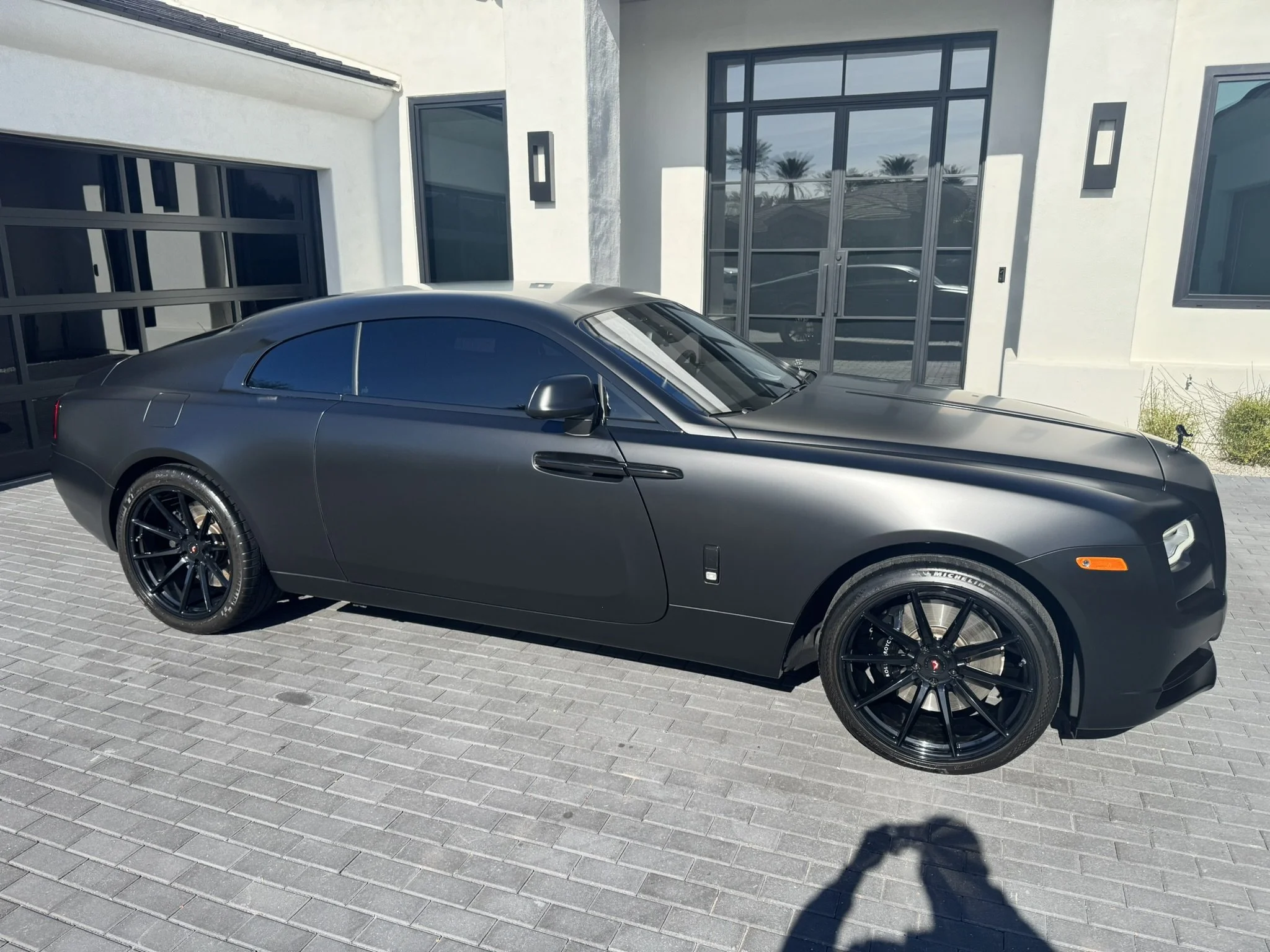 A black luxury coupe parked in front of a modern white house with large glass doors and windows.