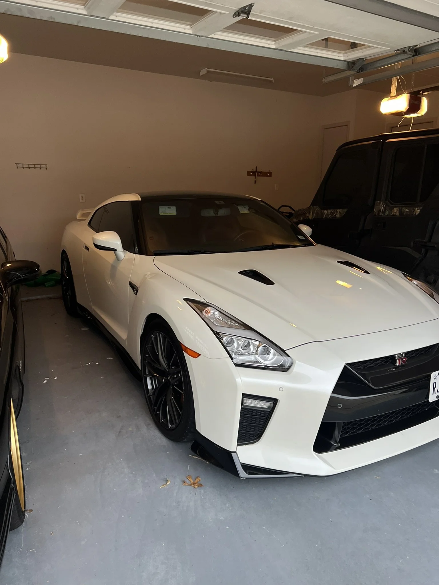 White Nissan GT-R sports car parked in a garage next to a black vehicle, with a beige wall in the background and a partially open garage door overhead.
