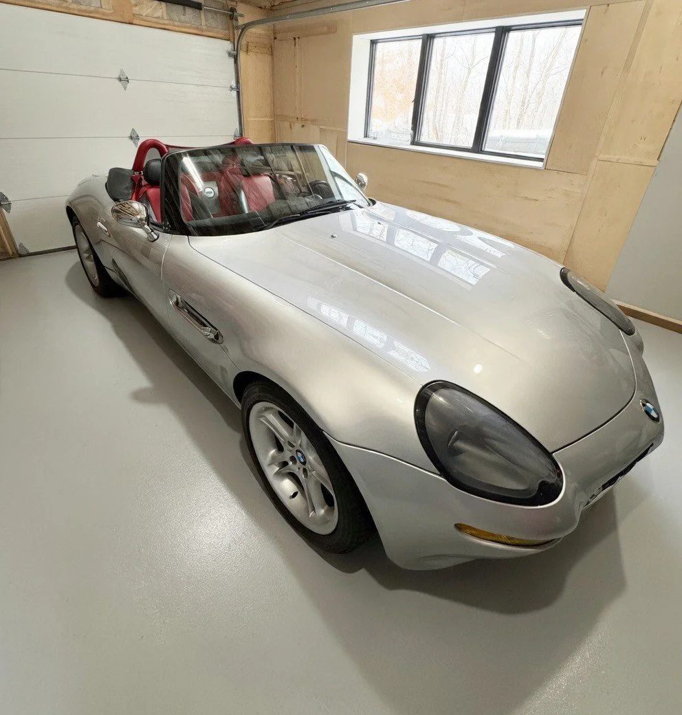 A silver BMW Z8 convertible with a red interior parked inside a garage, with a window on the side wall and a wooden wall paneling.