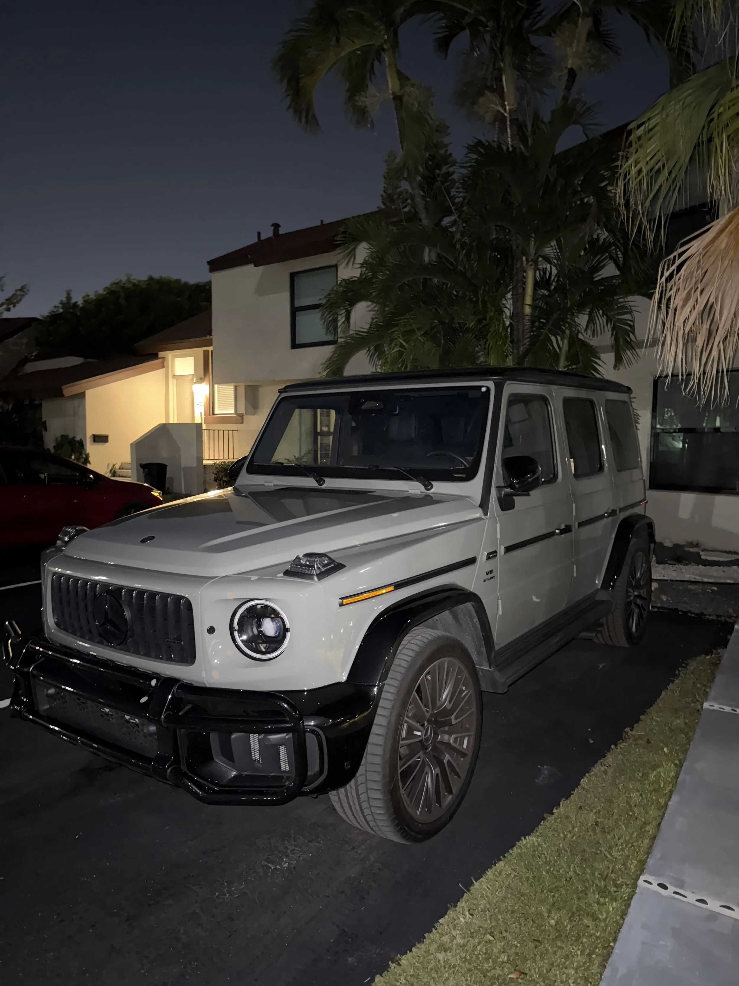 A white Mercedes-Benz G-Class SUV parked on a driveway at night, with a residential house, palm trees, and a neighboring vehicle visible in the background.