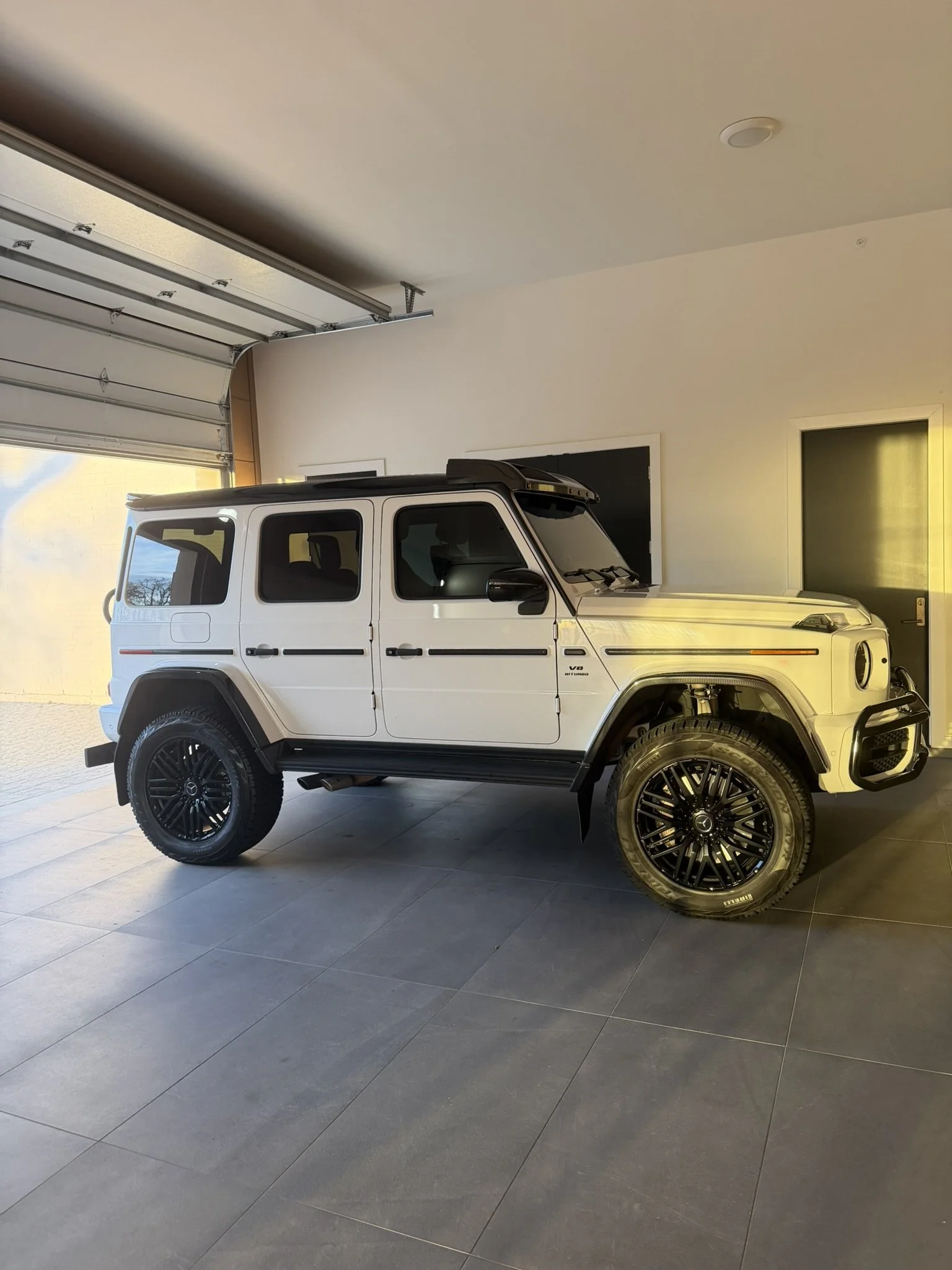 White Mercedes-Benz G-Class SUV parked inside a modern house garage with an open garage door, black wheels, and tinted windows.