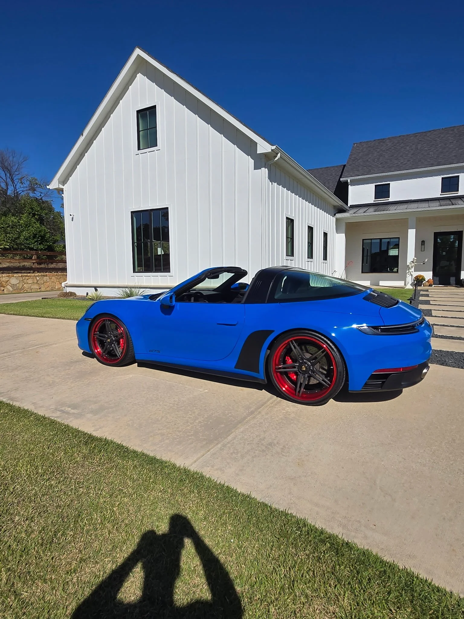A blue Porsche sports car with red wheels parked in front of a modern white house on a sunny day.