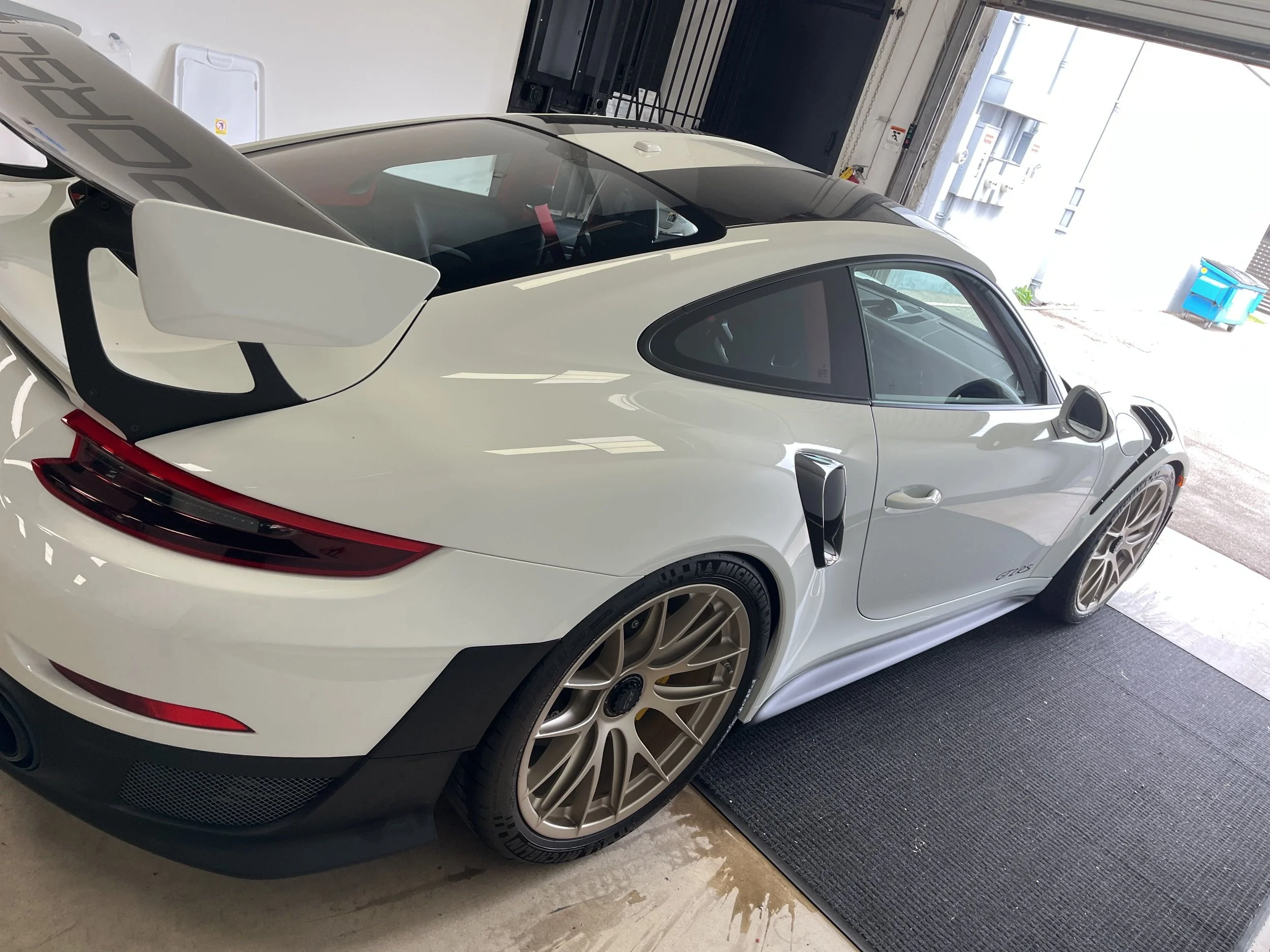 A white sports car with a large rear wing, gold rims, and racing modifications parked inside a garage.