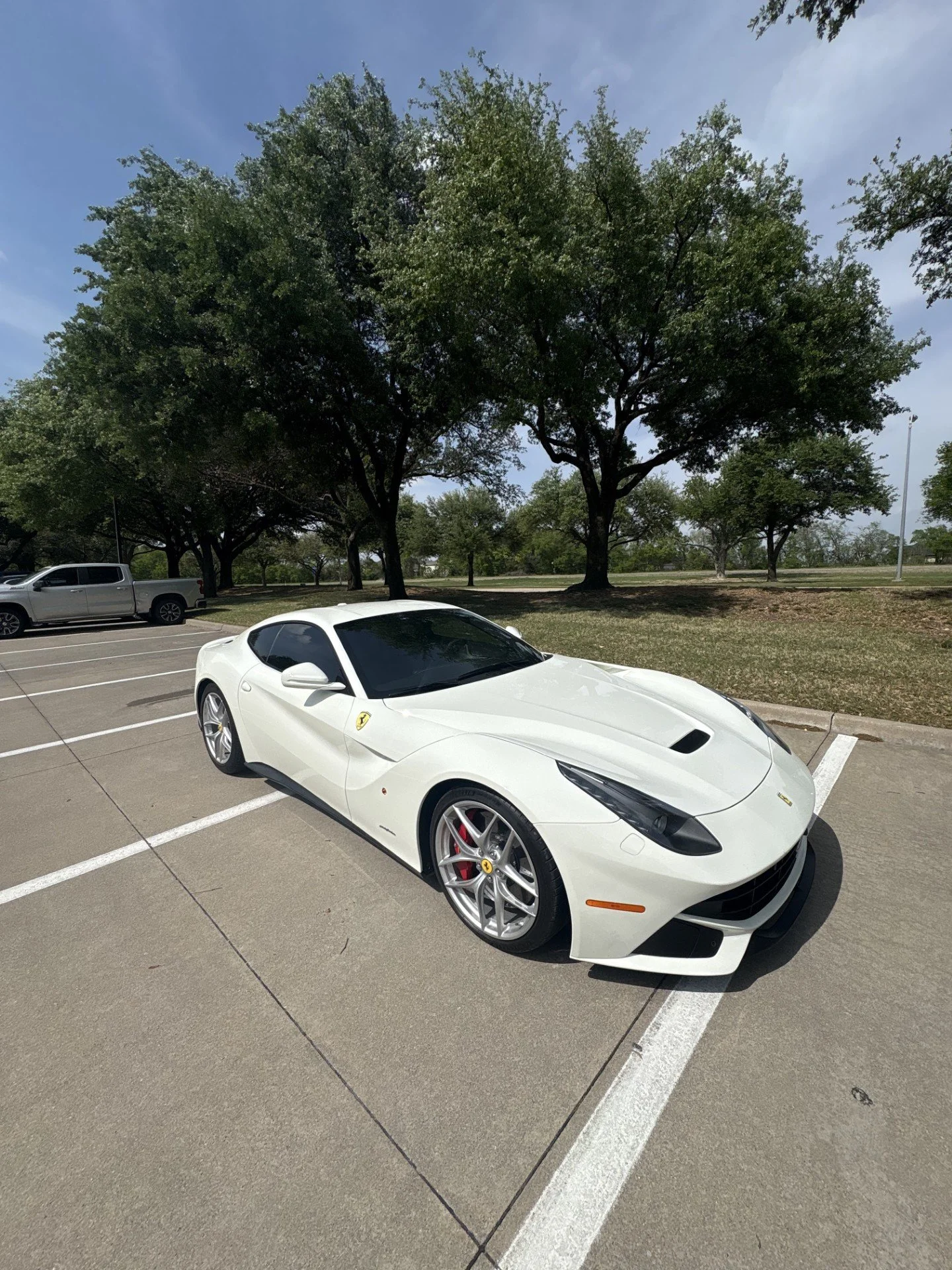 White Ferrari sports car parked in a parking lot, surrounded by trees and other vehicles under a partly cloudy sky.