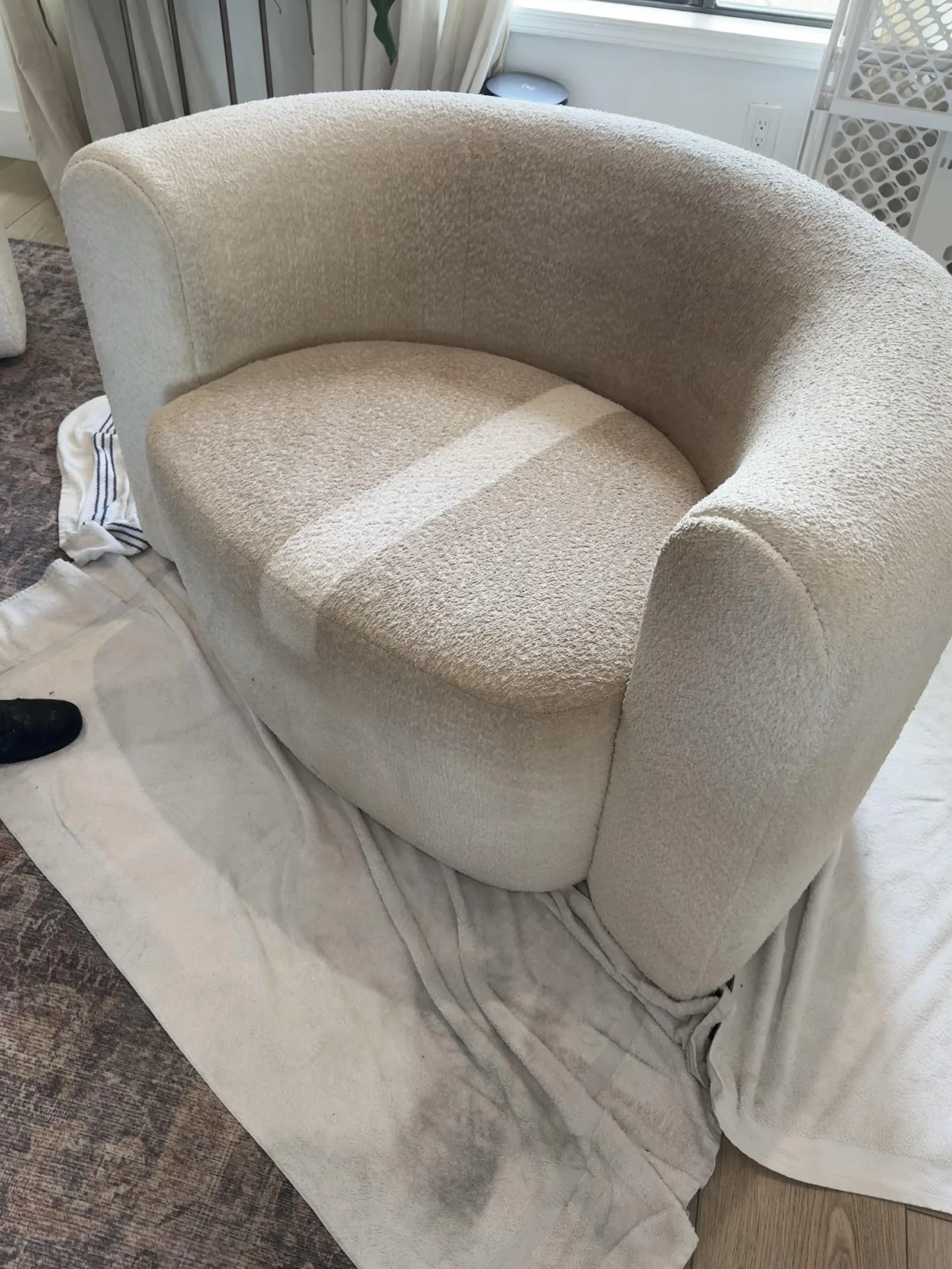A beige upholstered armchair with a rounded back and curved armrests. It is placed on a white sheet, with a floor visible underneath. The chair has one cleaned streak through the middle. Showing the difference made by cleaning.