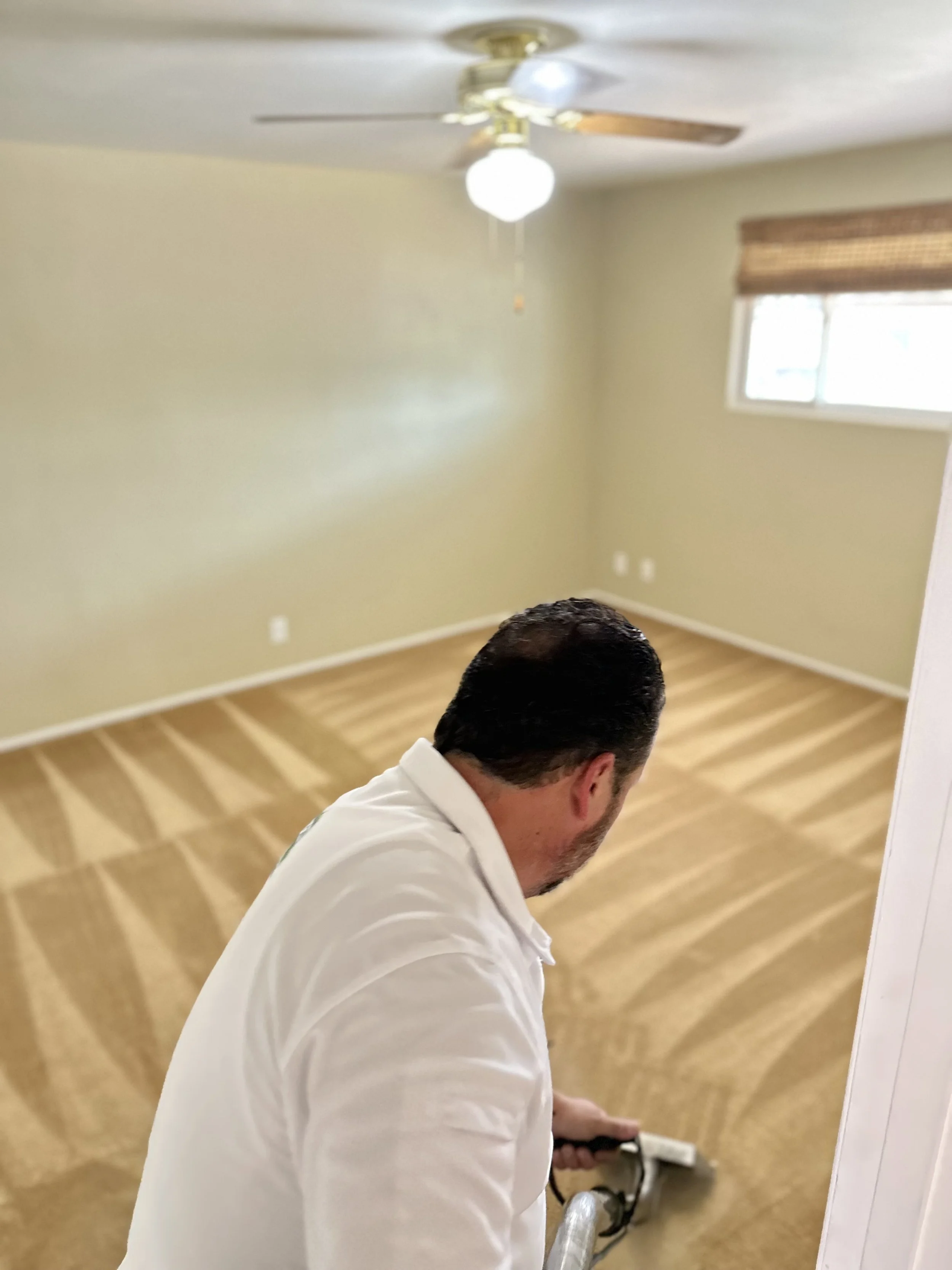 Man in white shirt cleaning carpet with a steam cleaner in an empty, beige-colored room with a ceiling fan and window.