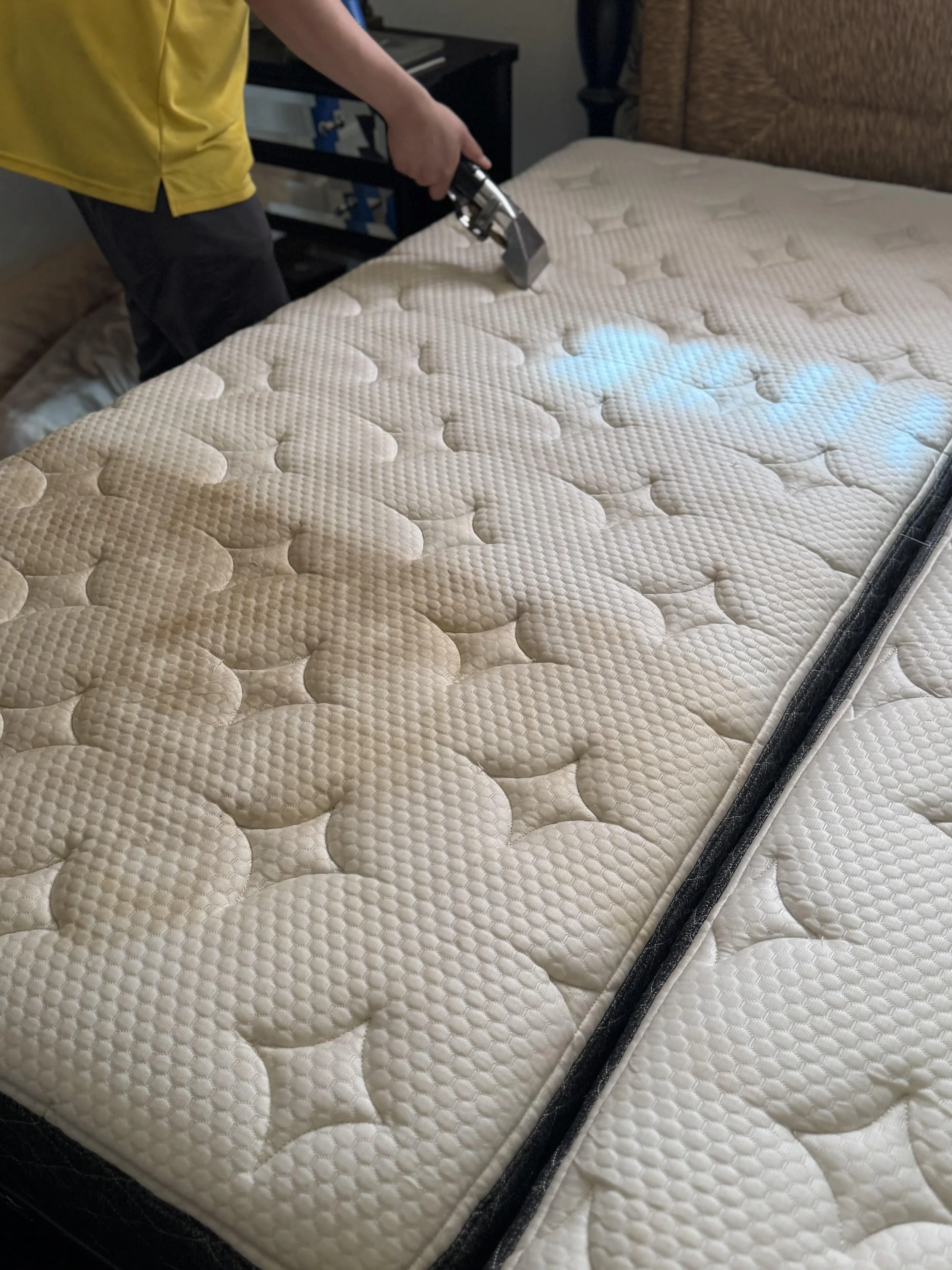 Person cleaning a mattress with a handheld steam cleaner in a bedroom.