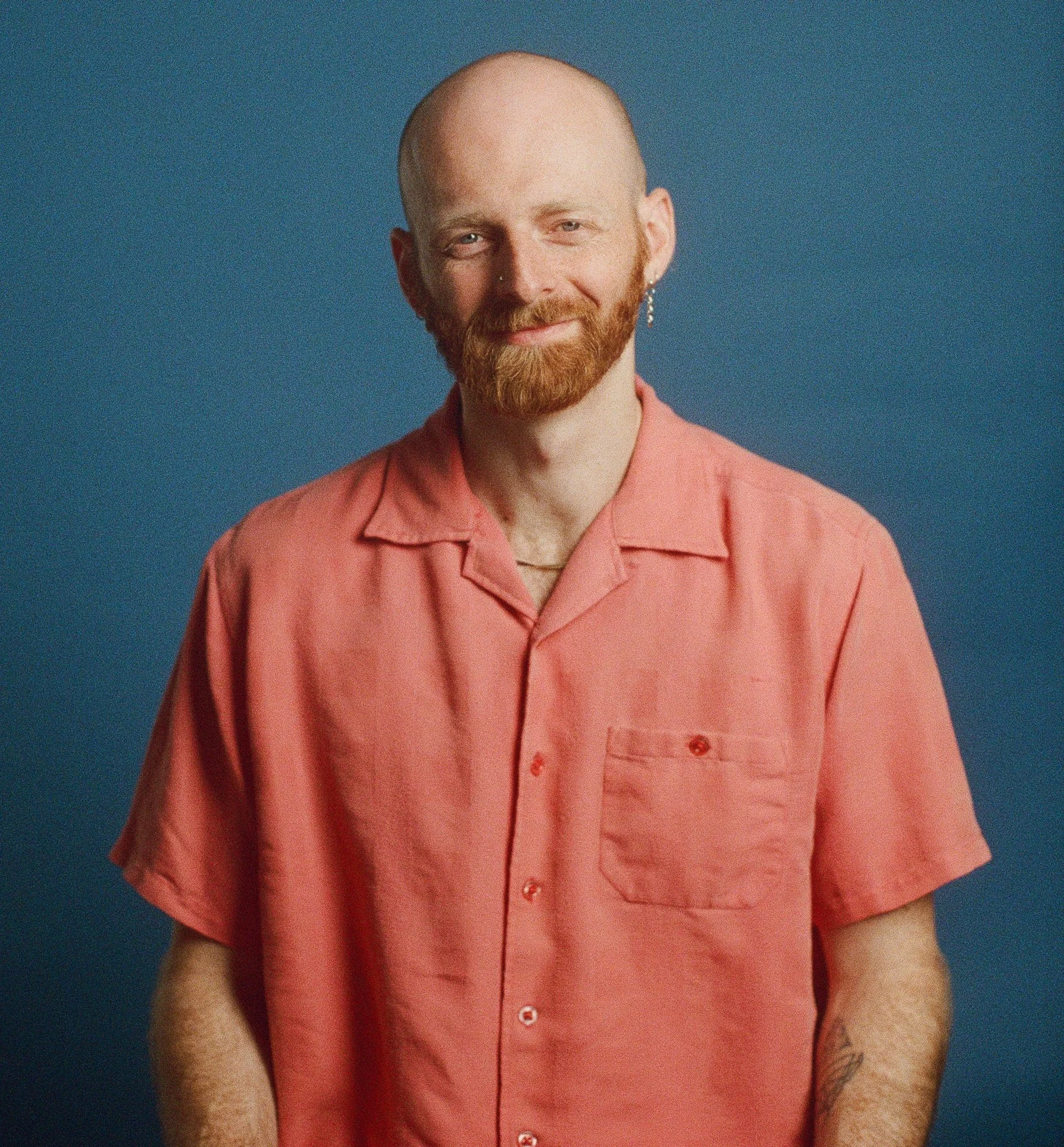 Close-up of a man with a shaved head, red beard, wearing a pink short-sleeved button-up shirt, earring, nose piercing, smiling, against a solid blue background.