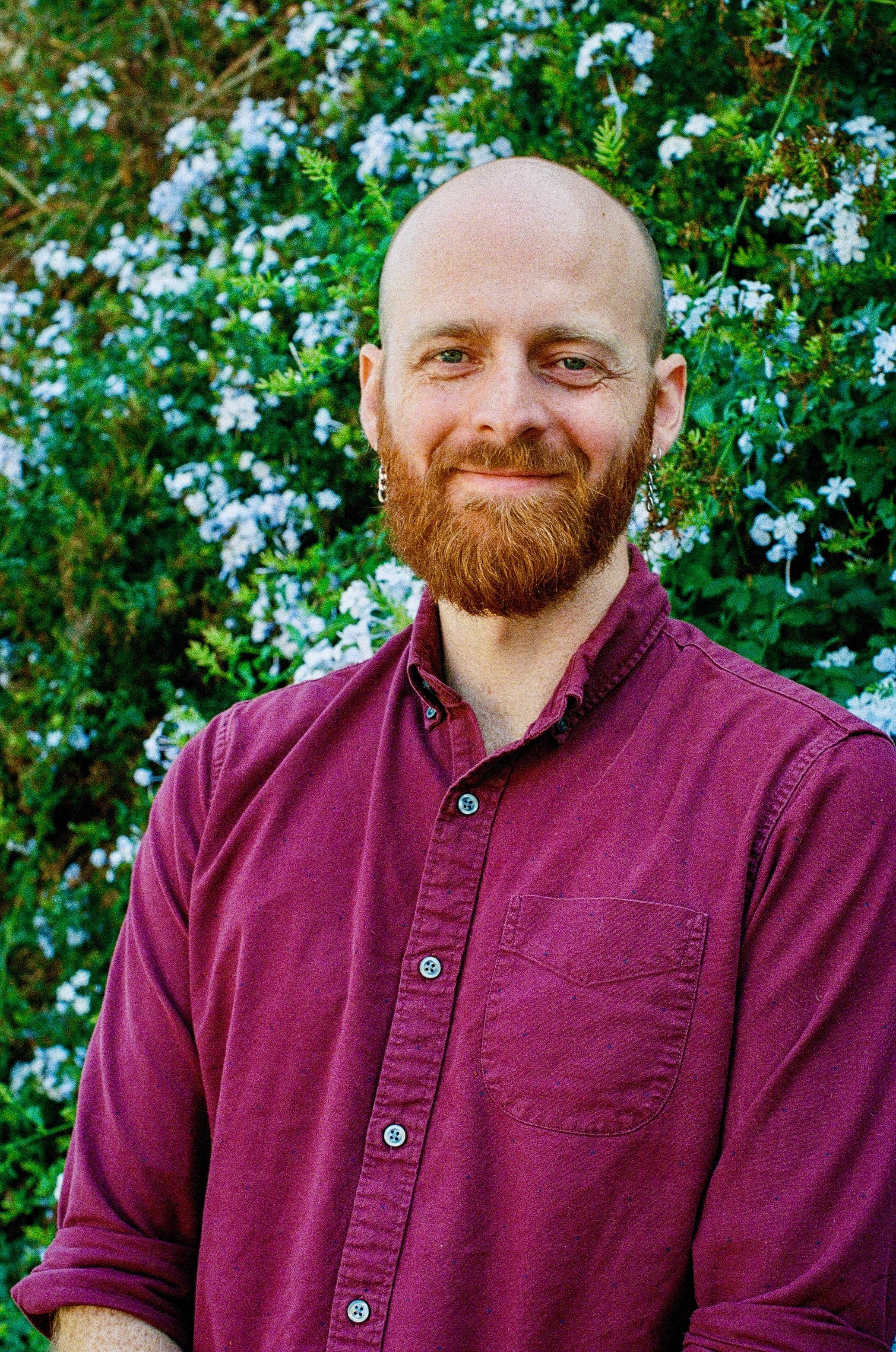 A man with a beard, wearing a maroon button-up shirt, smiling, standing in front of a bush with small white flowers.