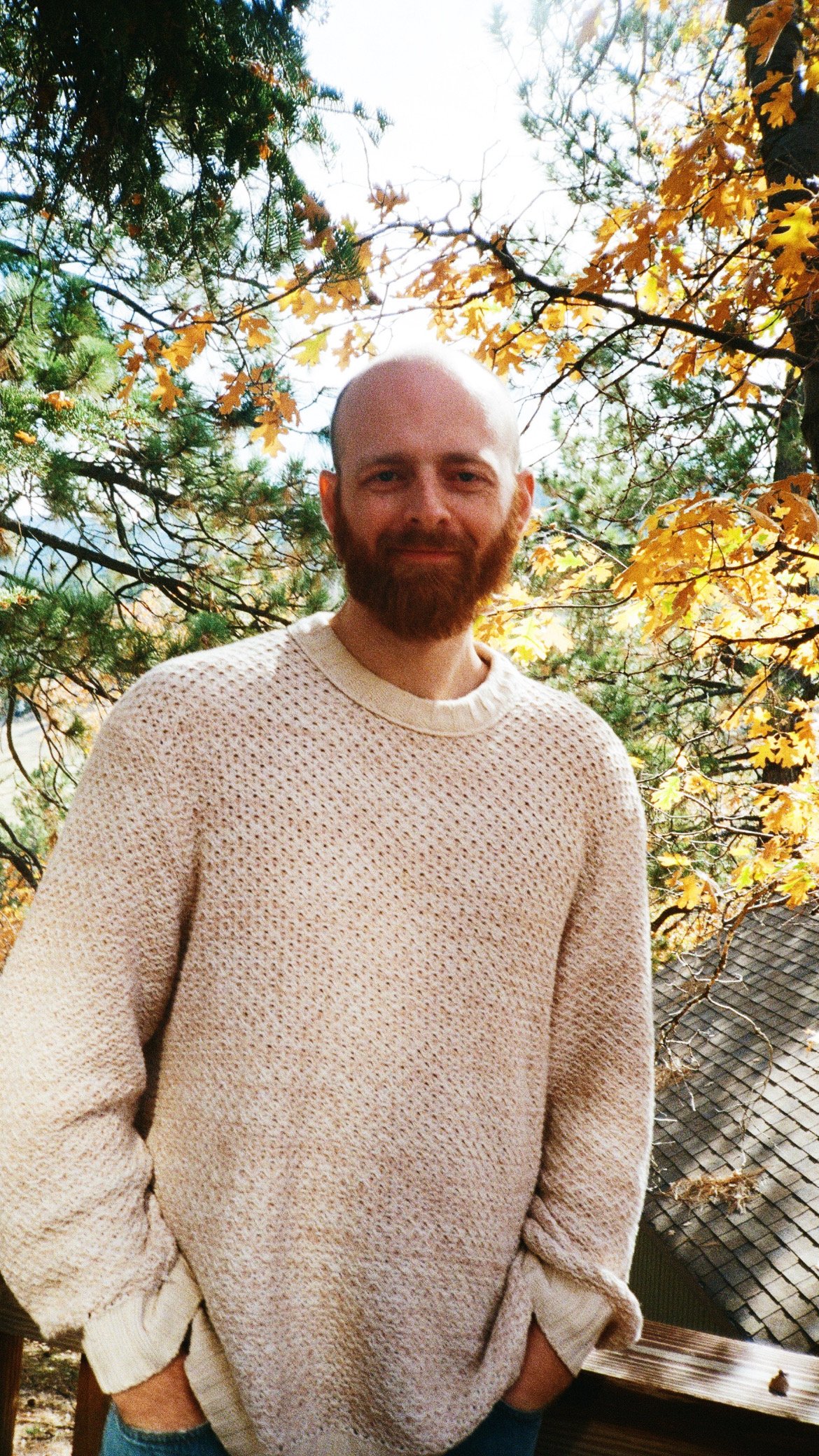 A man with a beard and a bald head relaxed outdoors on a sunny day, wearing a cream-colored knit sweater with trees and autumn leaves in the background.