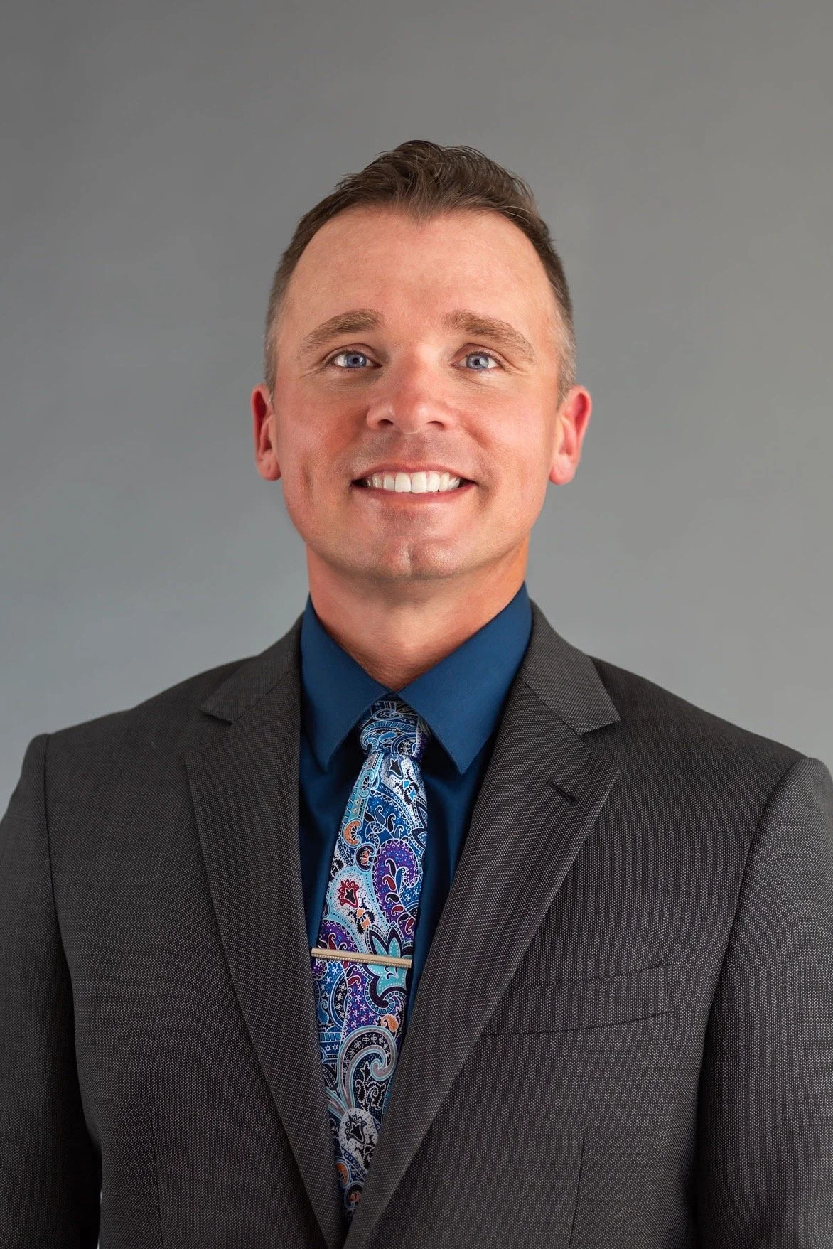 Professional portrait of a smiling man in a gray suit, blue shirt, and colorful paisley tie against a neutral gray background.