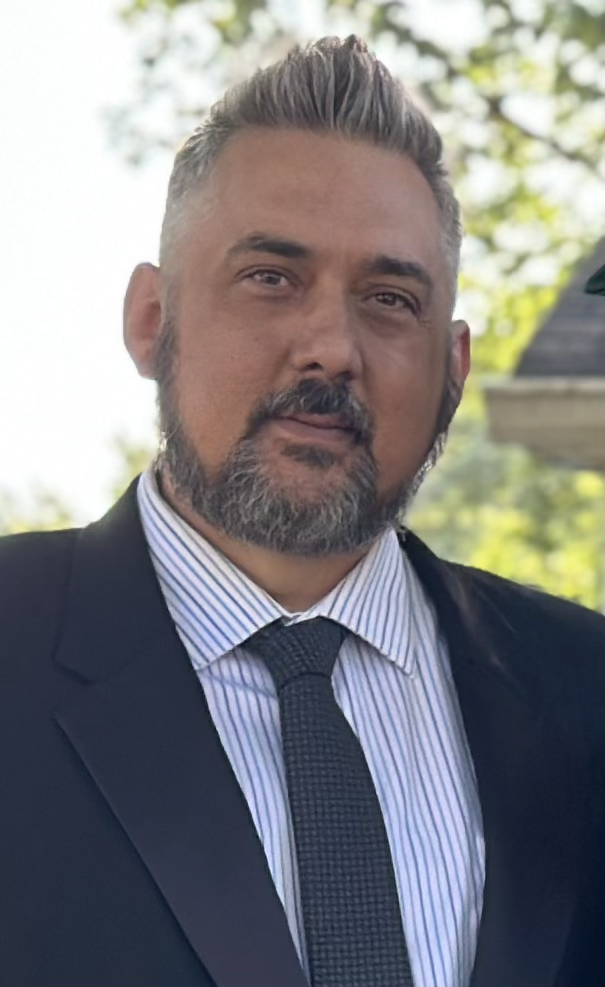 A man in a dark suit, with a striped dress shirt and a dark tie, standing outdoors with trees in the background.