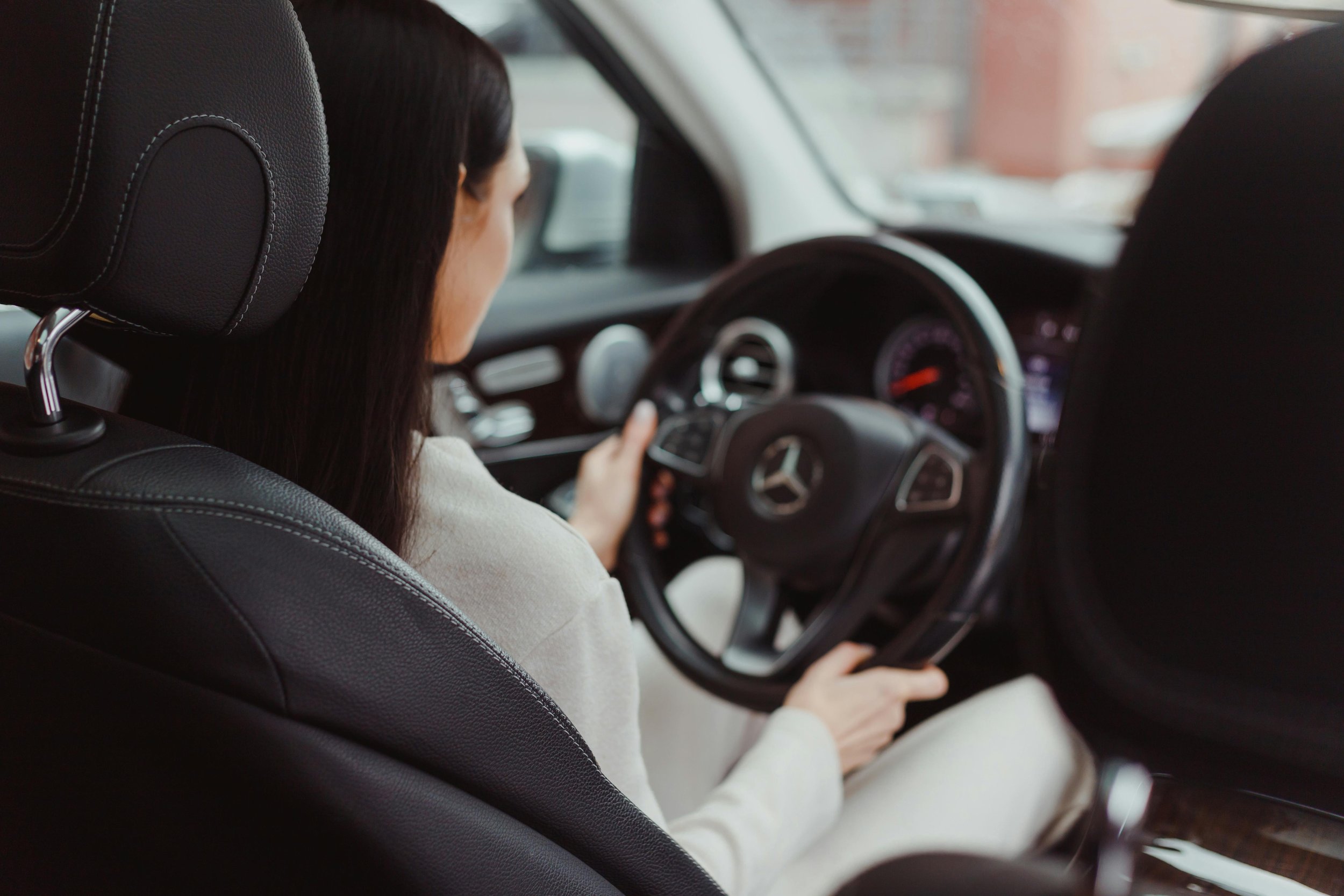 A woman driving a black Mercedes-Benz vehicle, viewed from the back seat, with focus on the steering wheel and dashboard.