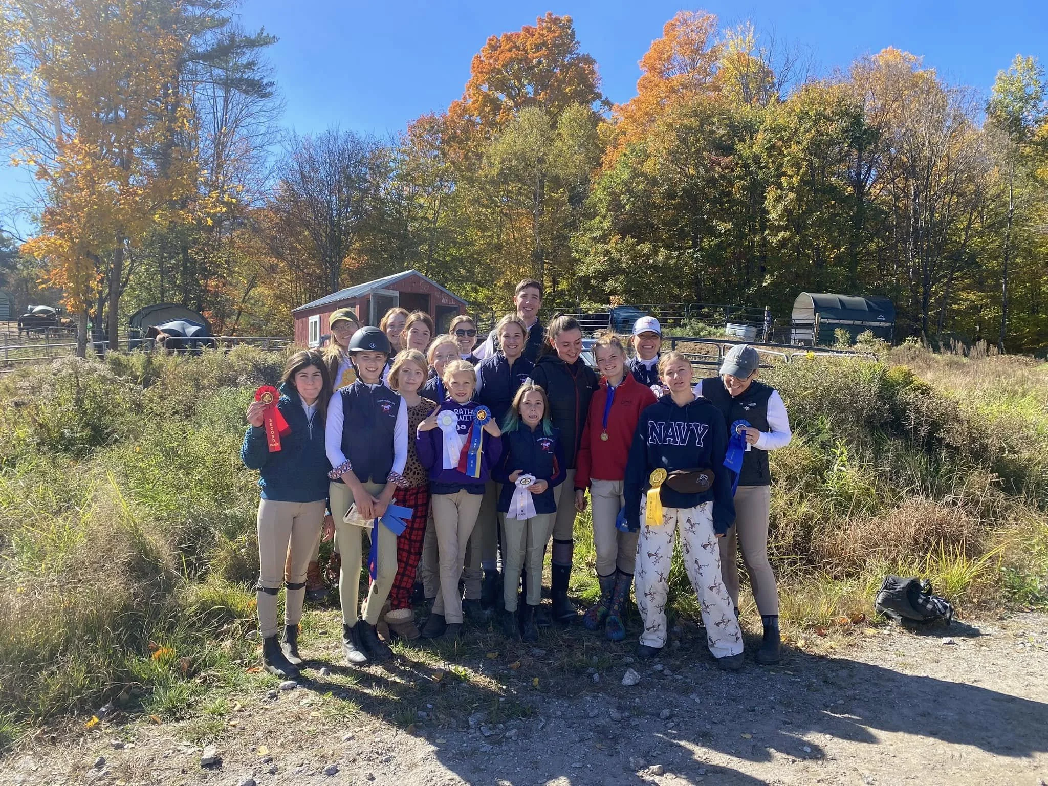 Group of young girls and women, some wearing medals and ribbons, standing outdoors on a sunny day with autumn trees in the background.