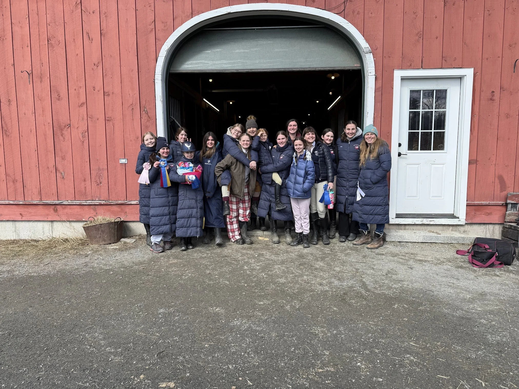 Group of people, mostly children wearing winter coats, standing outside a red barn with a white door, smiling for a photo.